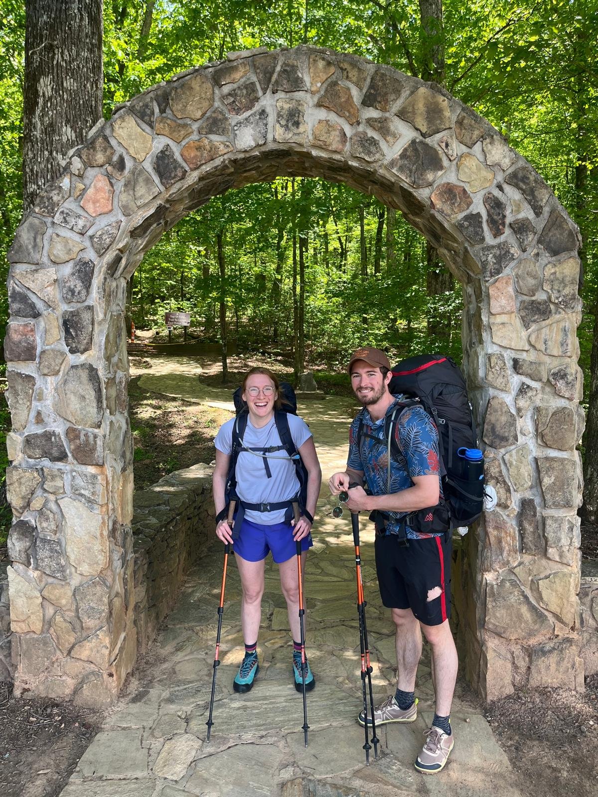 A smiling young woman and man with backpacks and hiking poles standing under a stone archway in a forest.