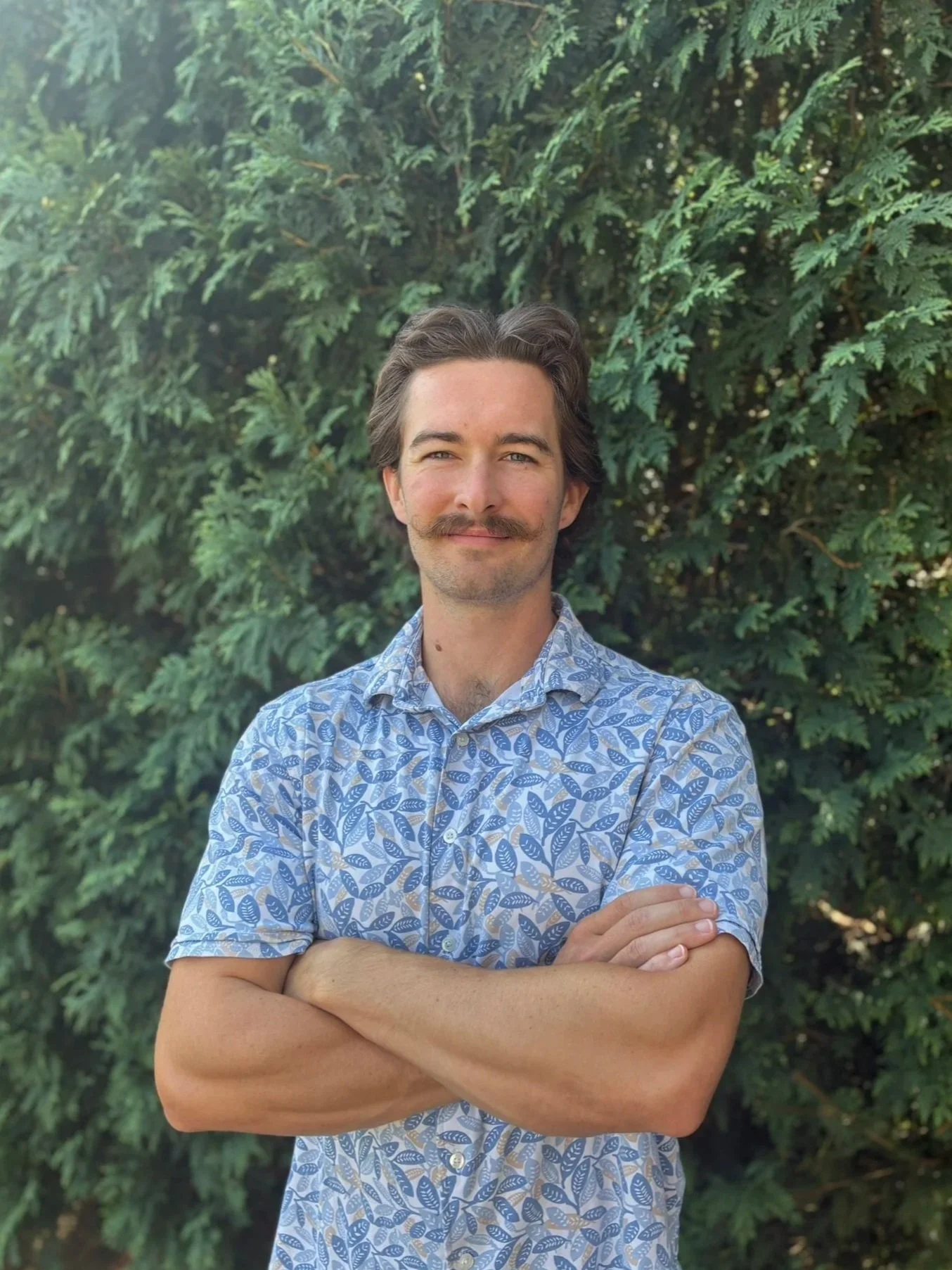A man with dark hair, a mustache, and a light complexion standing outdoors in front of a large, green, leafy shrub. He is wearing a short-sleeved, button-up shirt with a blue and white leaf pattern. He has his arms crossed and is smiling slightly at the camera.