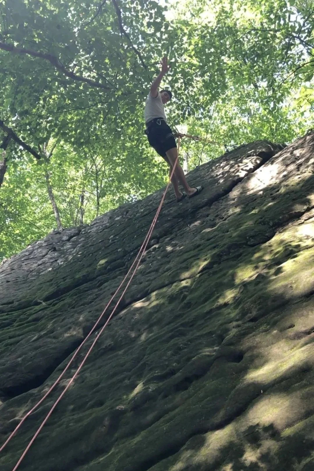 A coach rock climbing a steep rock wall in a forested area, using a harness and safety ropes.