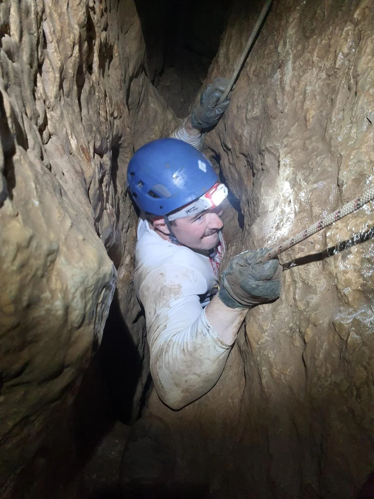 A man wearing a blue helmet, gloves, and a dirty white shirt is crawling through a narrow, muddy cave, holding a rope for support.