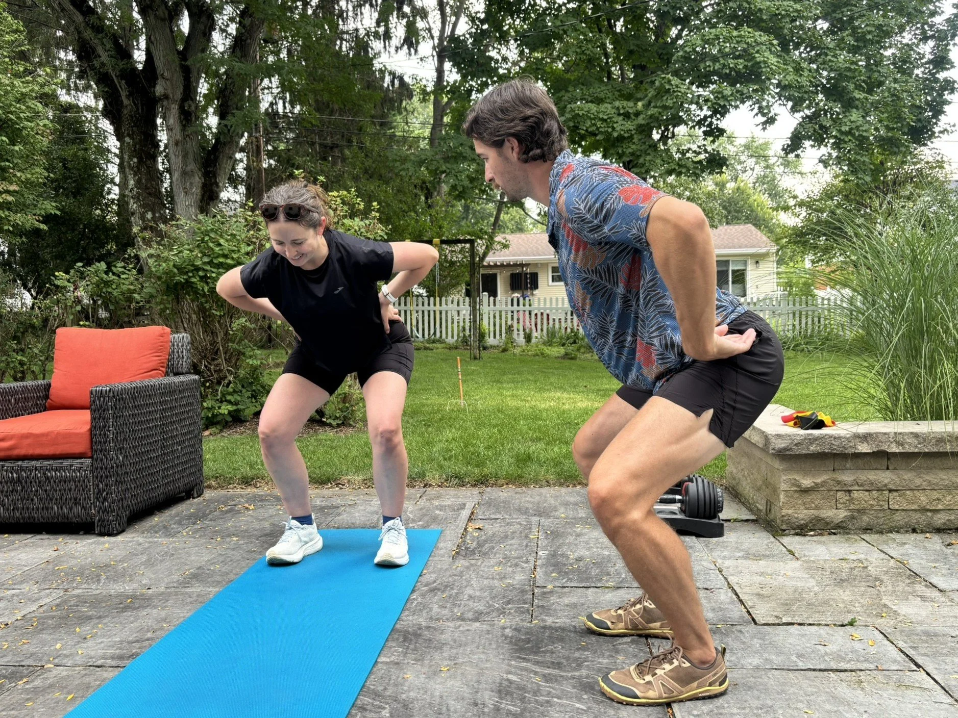 A man and a woman are exercising outdoors on a patio. They are in a squatting position, facing each other and smiling. The woman is wearing black athletic clothing and sunglasses on her head, and the man is wearing a patterned short-sleeve shirt and shorts. A blue exercise mat is beneath them. There is a black wicker chair with an orange cushion, some garden tools, and a grill nearby, with a house and trees in the background.
