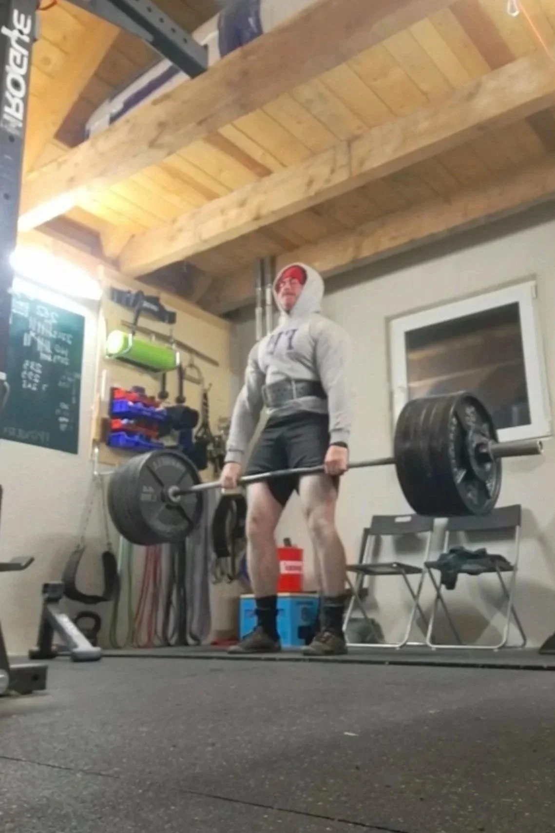 A personal trainer in a hoodie and shorts lifting (deadlifting) a heavily loaded barbell inside a gym.