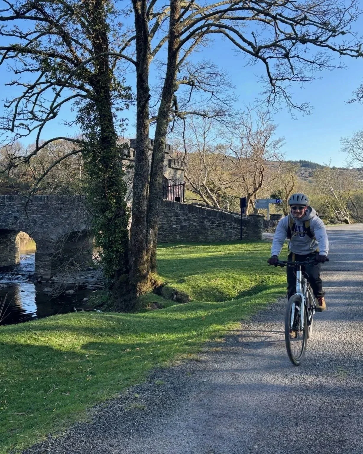 A personal trainer riding a bicycle (cycling) on a trail beside a grassy area and a small stream, with a stone bridge and building in the background, trees with bare branches, and a blue sky.