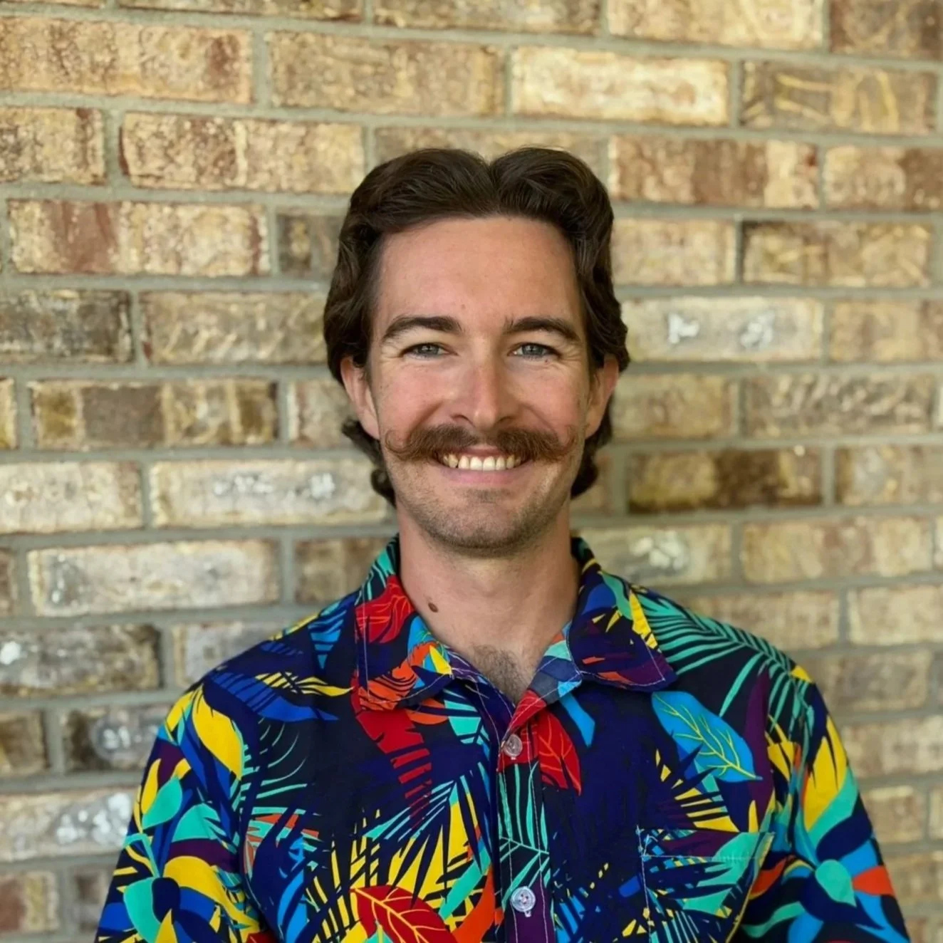 A coach with brown hair, a mustache, and a beard, smiling, standing in front of a brick wall, wearing a colorful tropical shirt.