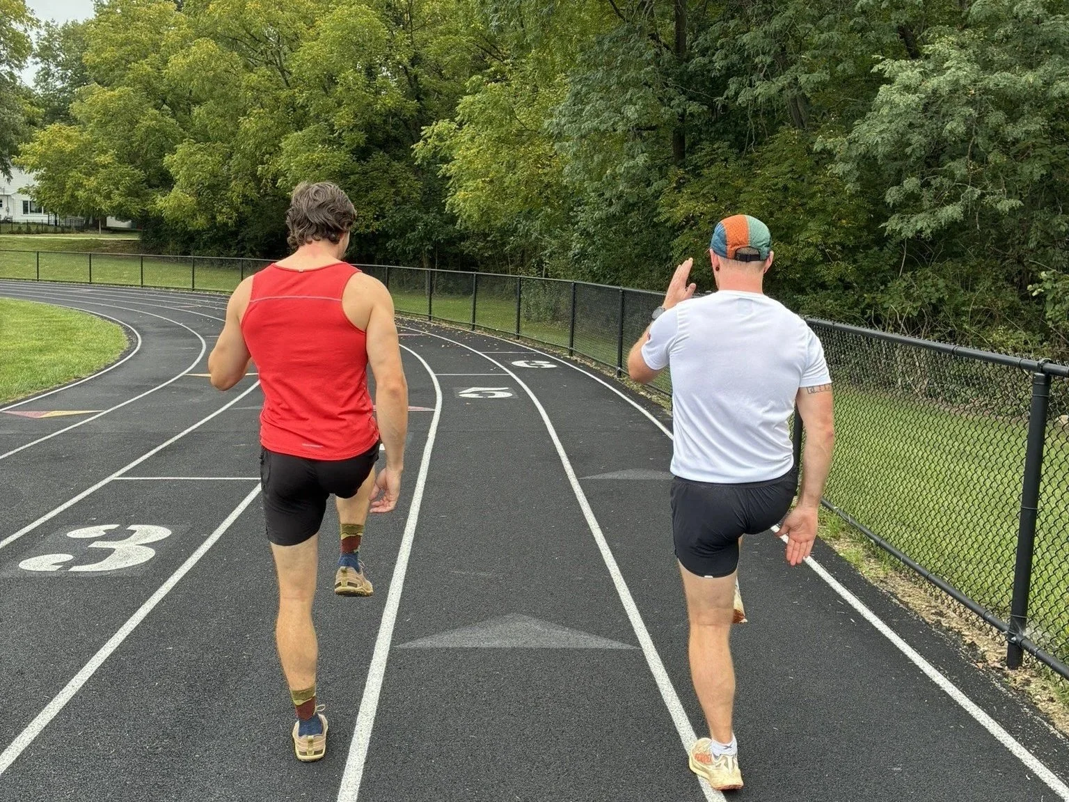 Two men running on a track surrounded by green trees, one wearing a red tank top and black shorts, and the other wearing a white T-shirt, black shorts, and a colorful cap.