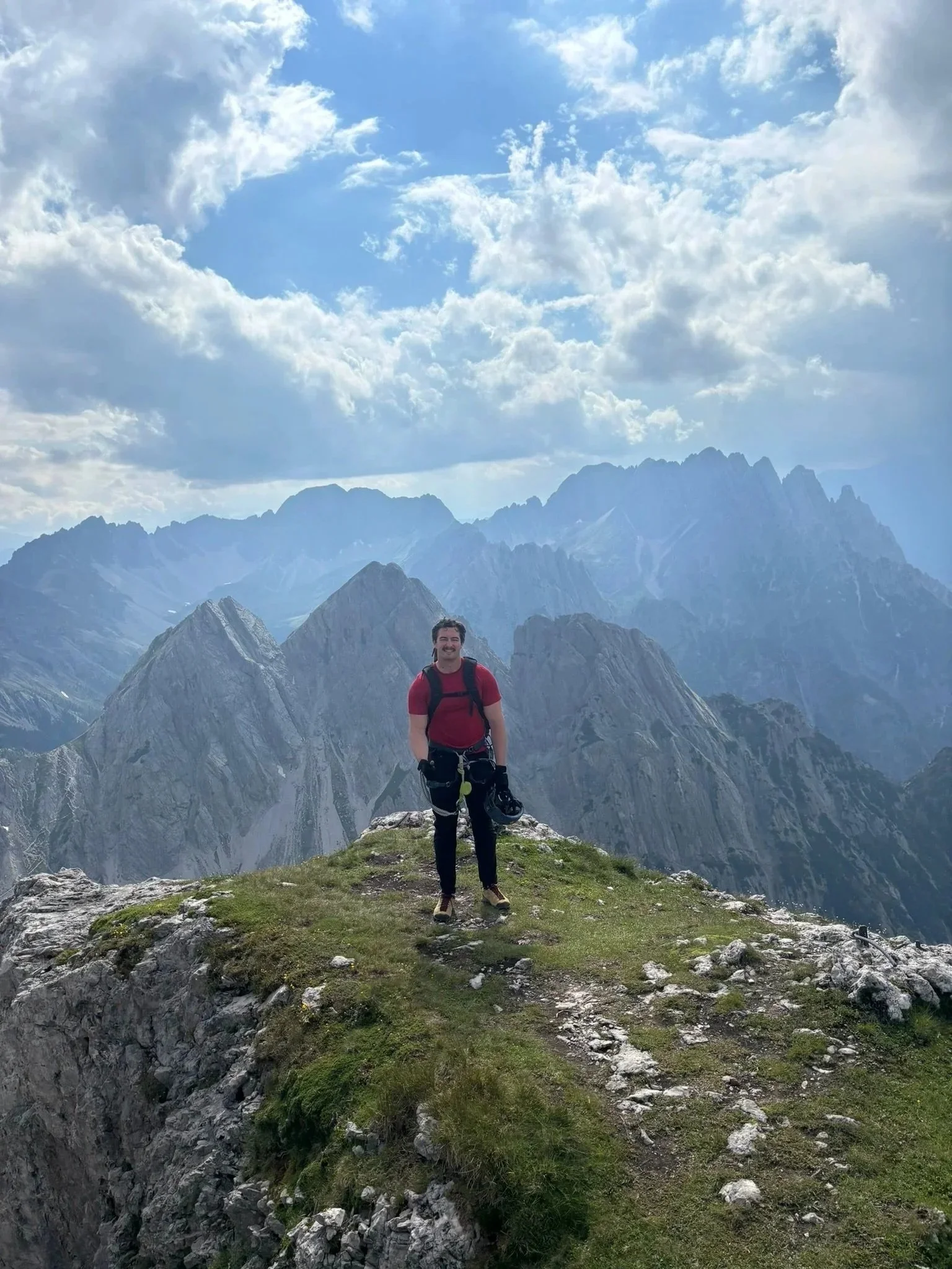 A coach standing on a grassy mountain peak with rugged mountain ranges in the background and a partly cloudy sky overhead.