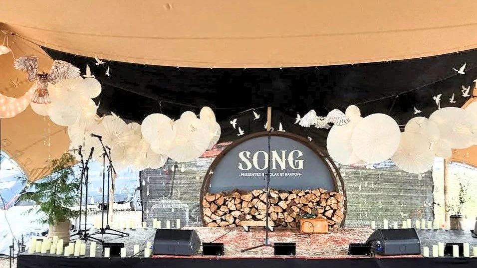 Decorated outdoor stage with white paper lanterns, artificial birds and trees, and a wood storage behind a sign reading 'SONG' for a musical event. Owl sculptures created by Harlequin arts hang from the stage.