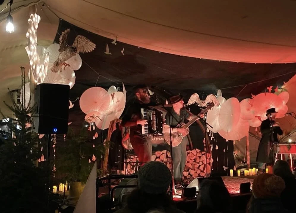 Musicians performing on a decorated stage with paper lanterns and hanging art, in a dimly lit indoor or tented venue. The owl sculptures created by Shannon Moore and Harlequin Arts are hanging from the stage.