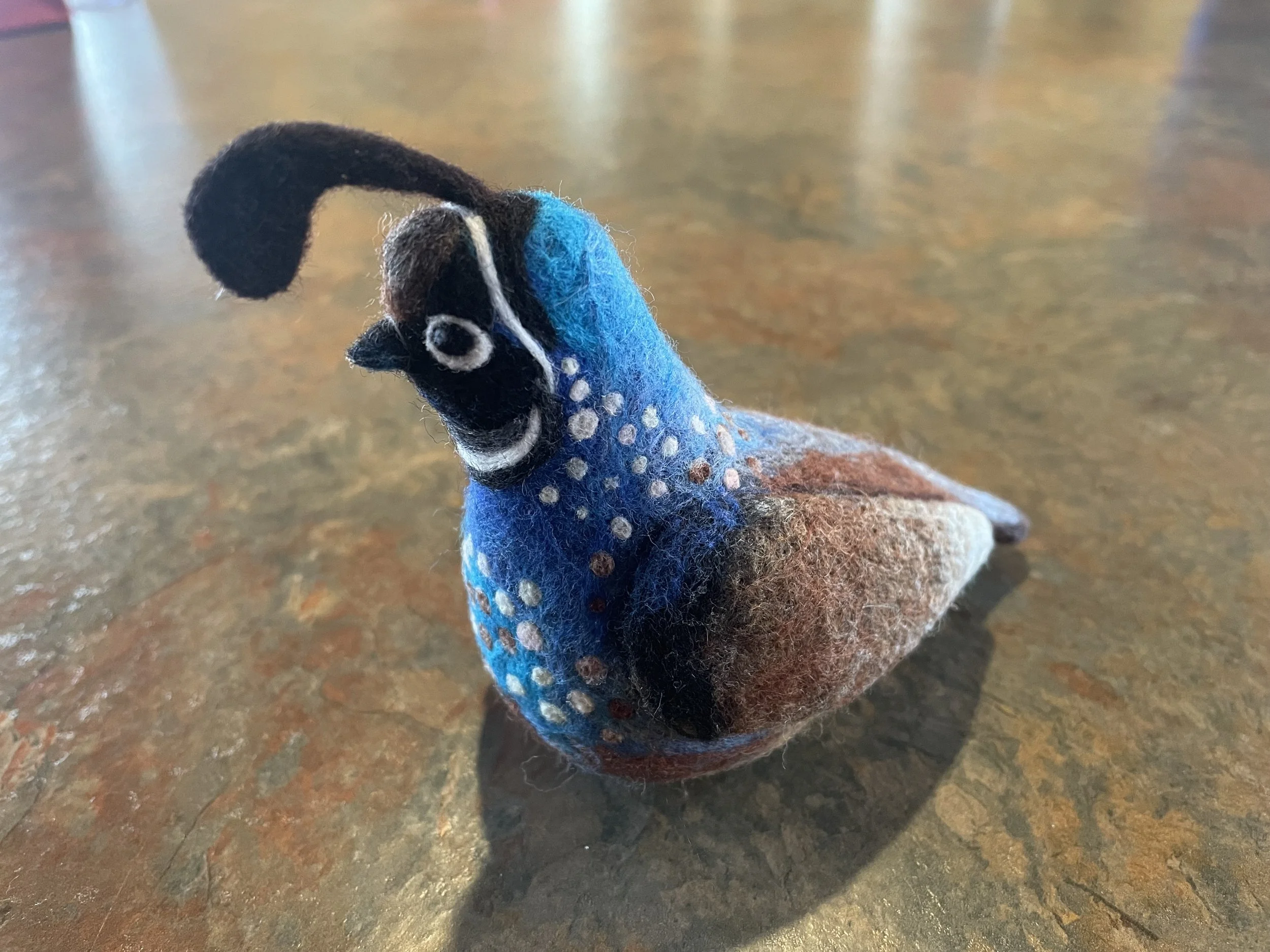 A felted animal figure of a blue pheasant with a black face, black crest, brown back and wings, and white spots all over.