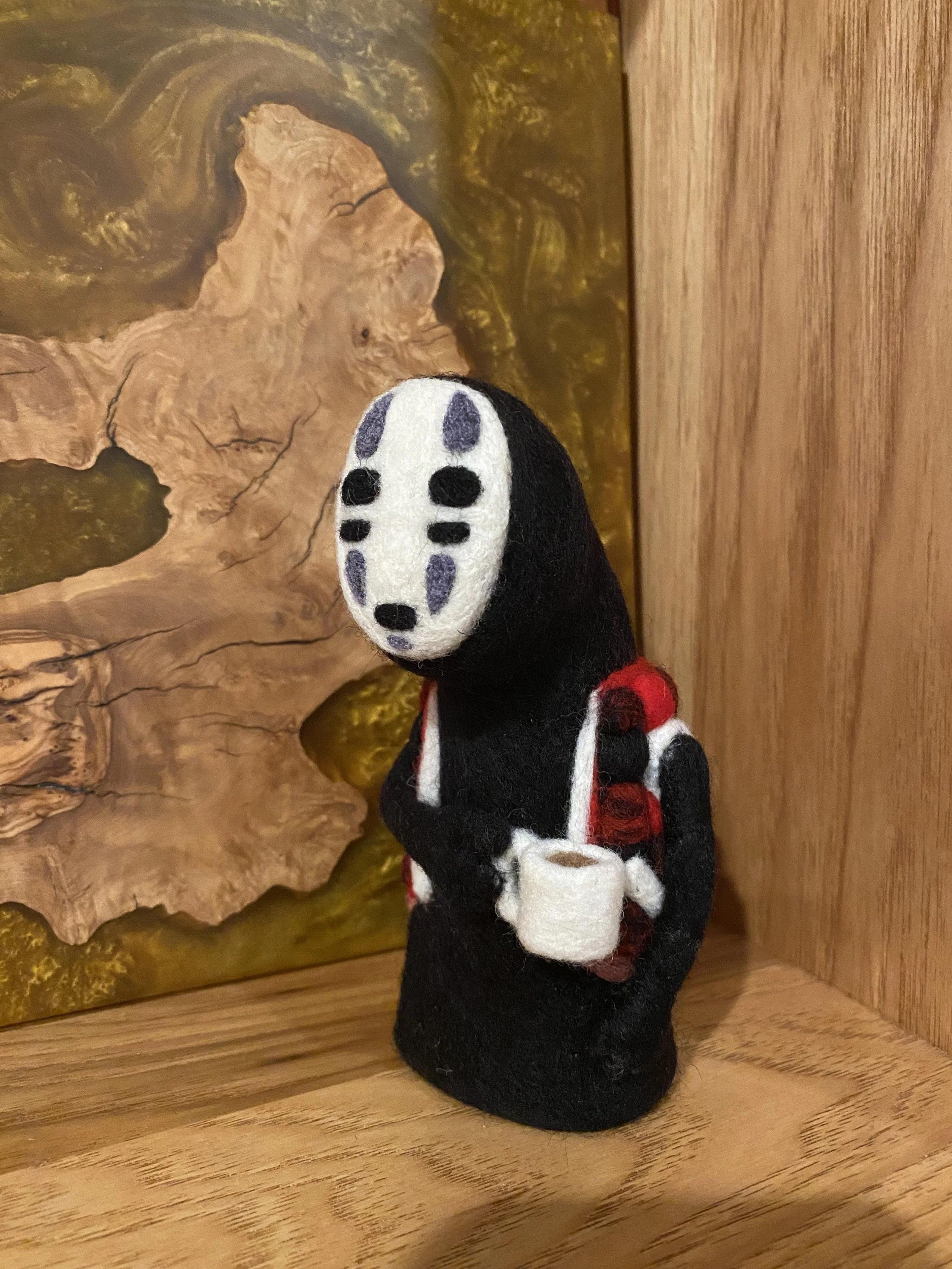 An 8 inch tall needle felted No-Face from Spirited Away that appears to be Pacific Northwest themed, wearing a red and black plaid vest, and holding a coffee cup in front of a wooden background.