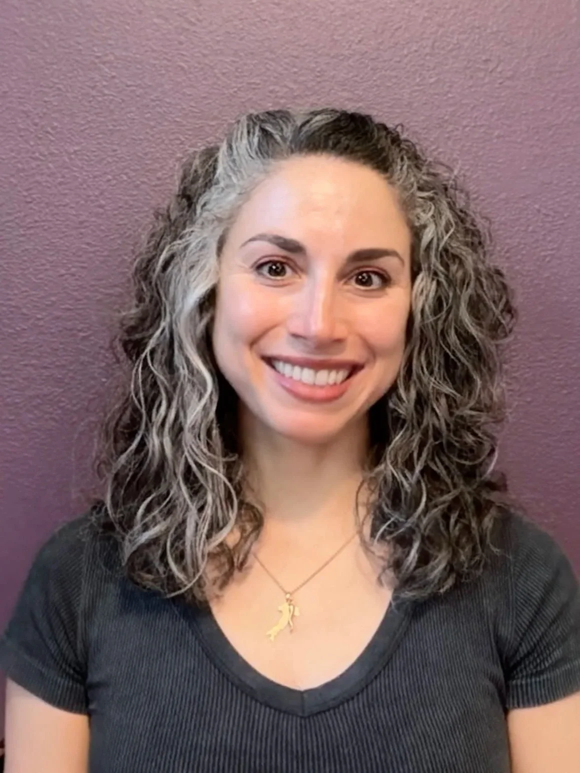 A woman with curly gray and brown hair smiling in front of a purple wall, wearing a black shirt and a gold necklace.
