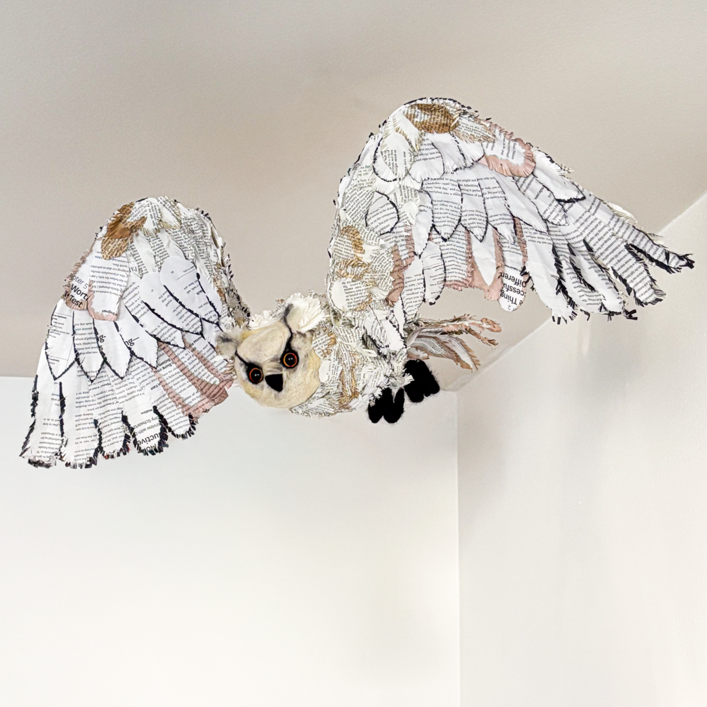 An owl sculpture with wings made of layered pages of text and a needle felted face and feet, hanging from white wall. Looks like a barn owl.