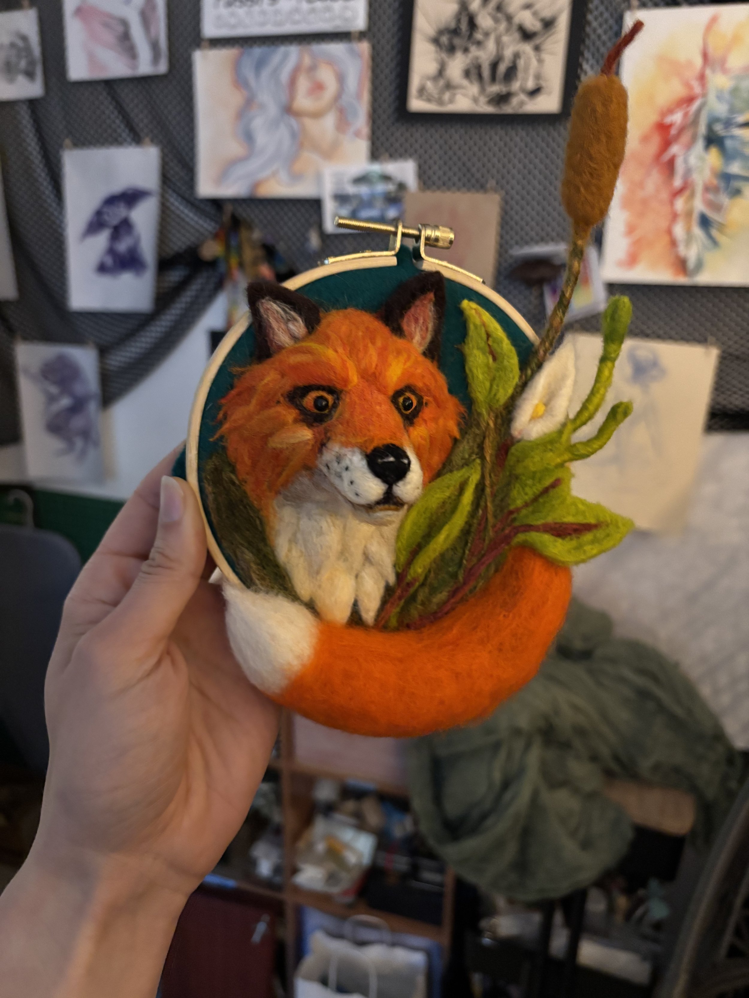 A needlepoint artwork of a fox's face and tail, with felted wool leaves, held by a Shannon Moore in an art studio with paintings on the wall.