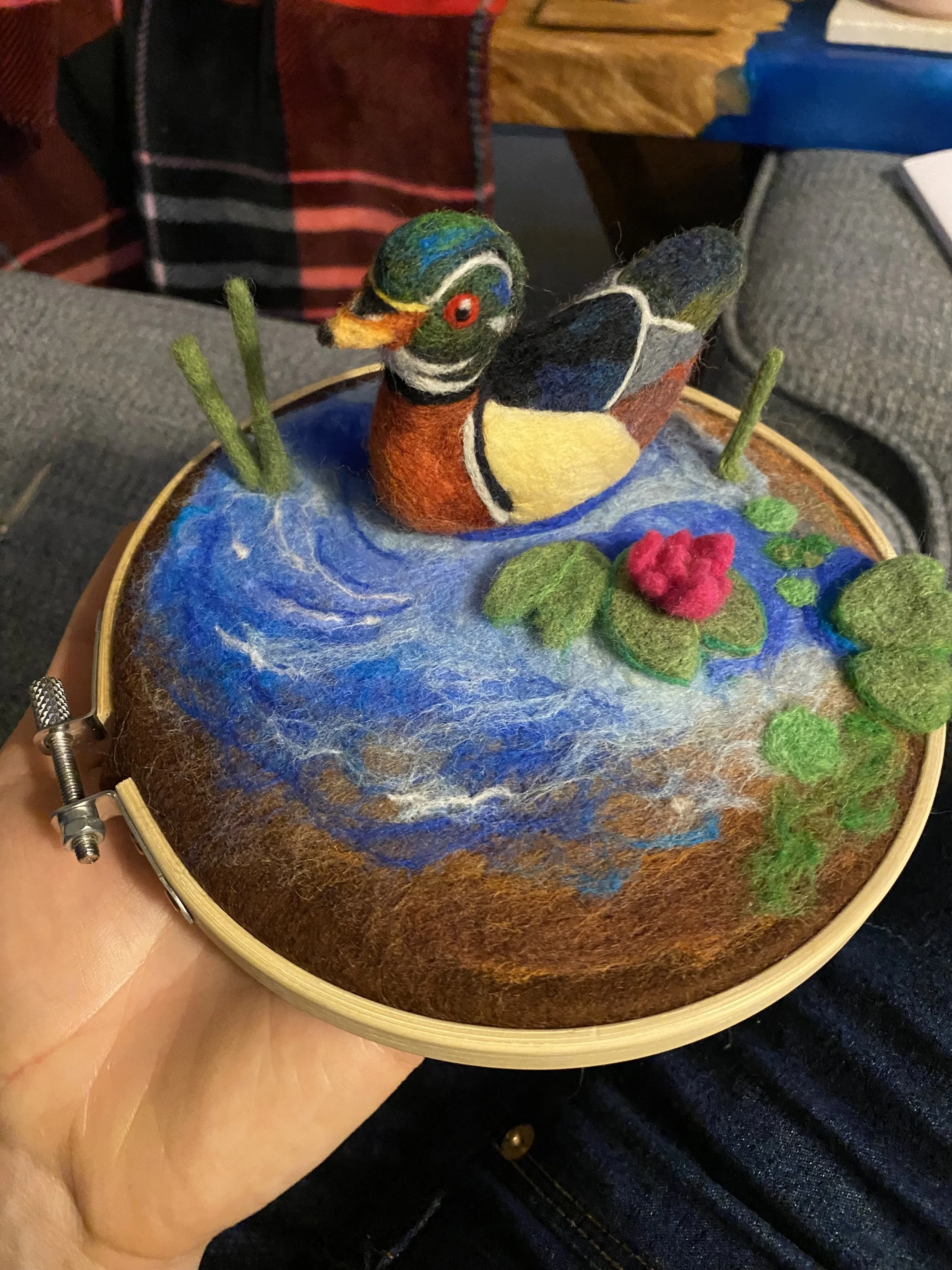 A handcrafted felted artwork of a duck resting on water, inside a embroidery hoop, with a lily pad and a pink water lily.