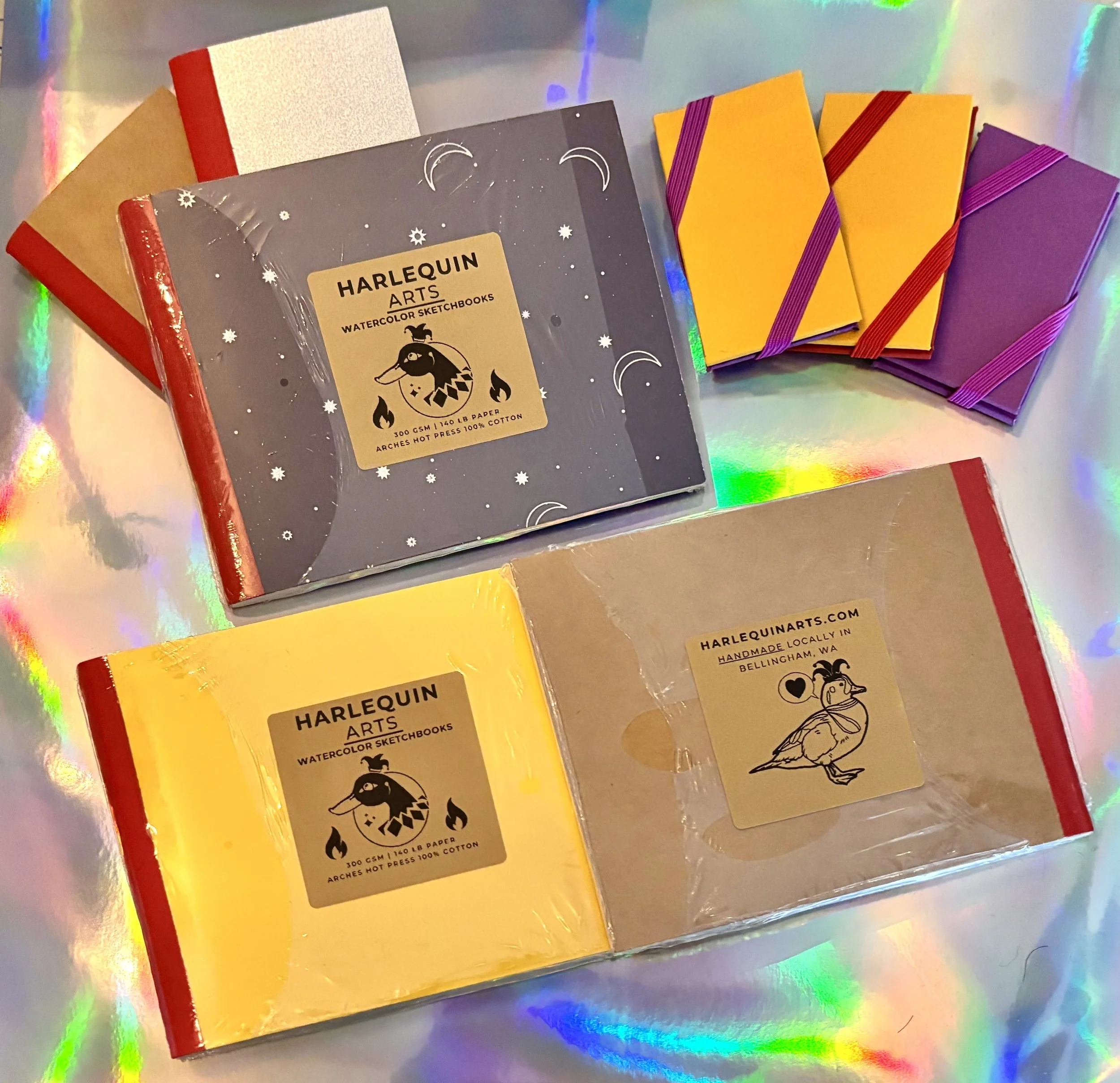 Various colorful hardcover sketchbooks with elastic bands, including a dark gray Harlequin Arts watercolor sketchbook in the center, on a holographic surface.