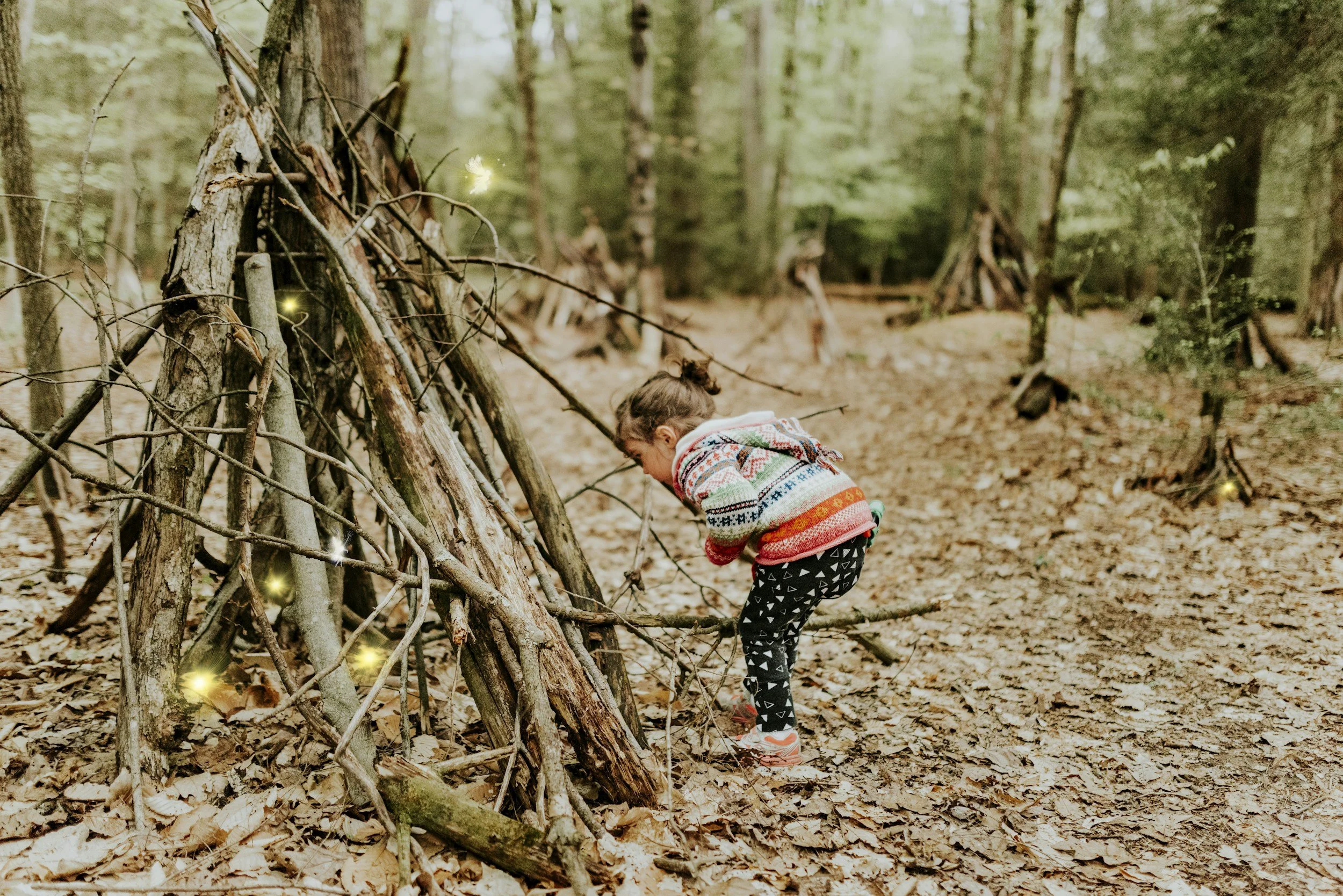 Niña con suéter colorido jugando en un bosque con estructuras de ramas y troncos, rodeada de hojas secas, este tipo de juego libre es el que buscamos en GUARIDA, una propuesta para cumpleaños infantiles diferentes en Santiago de Chile.