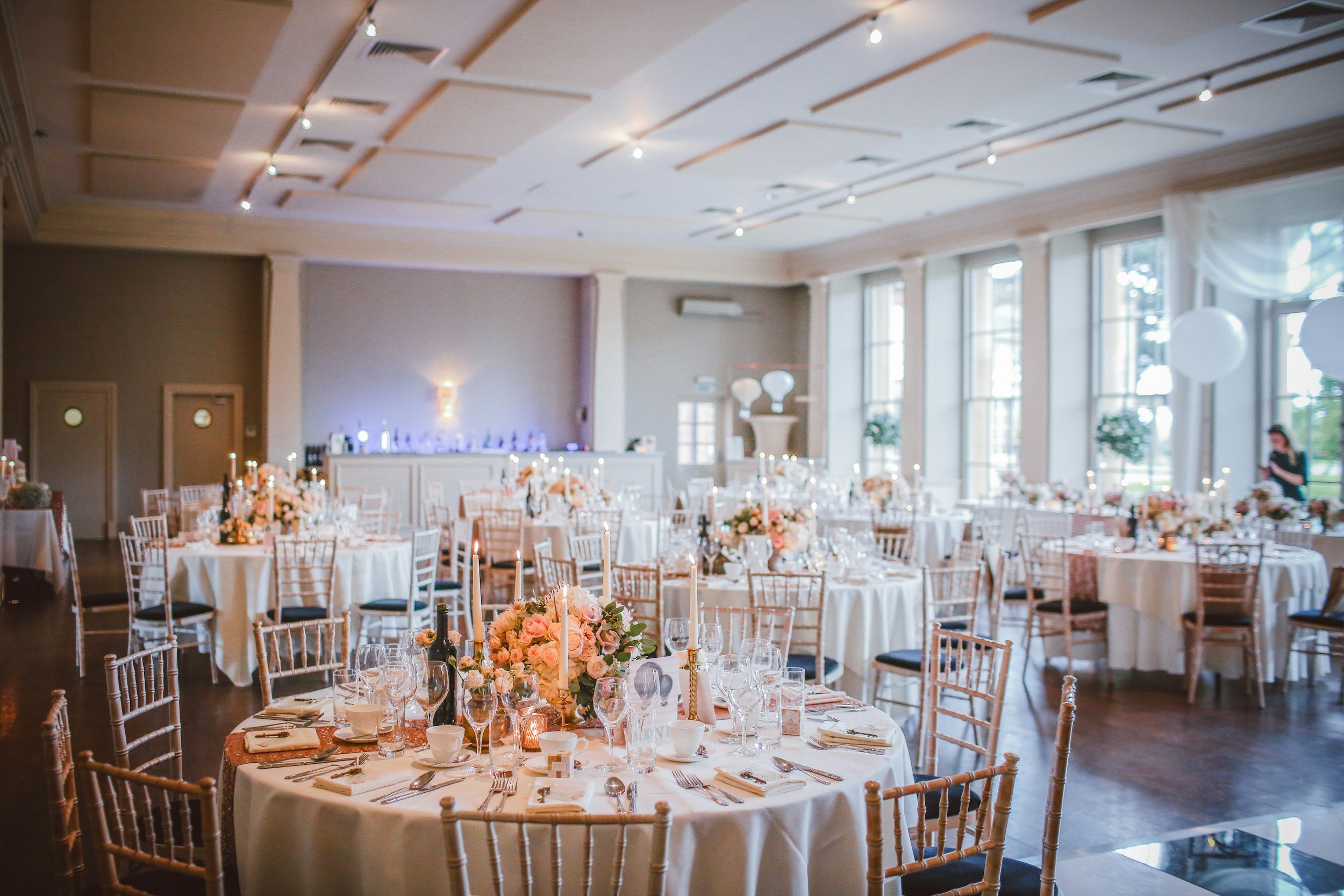 Elegant banquet hall decorated for a wedding reception with round tables covered in white tablecloths, set with glassware, cutlery, and floral centerpieces, surrounded by wooden chiavari chairs, with large windows allowing natural light, and soft lighting accents.