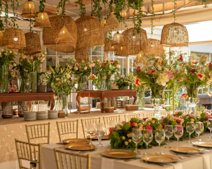 A beautifully decorated event space with hanging wicker light fixtures, floral centerpieces, and a long table set with plates, glasses, and gold-colored utensils for a celebration or wedding reception.