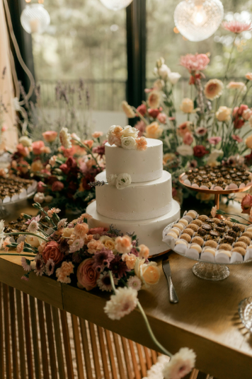 A two-tiered white wedding cake decorated with white flowers, surrounded by pink, peach, and cream-colored flowers on a wooden table with assorted desserts, set against large windows with greenery outside.