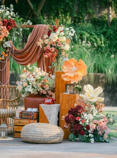 Bohemian outdoor wedding decor with floral arrangements, draped fabric, wooden accents, and seating on a wooden deck near a pond.