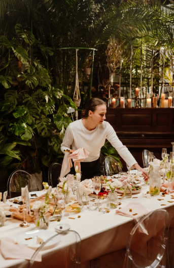 A woman setting a dinner table with candles and floral decorations in a lush, tropical indoor setting.