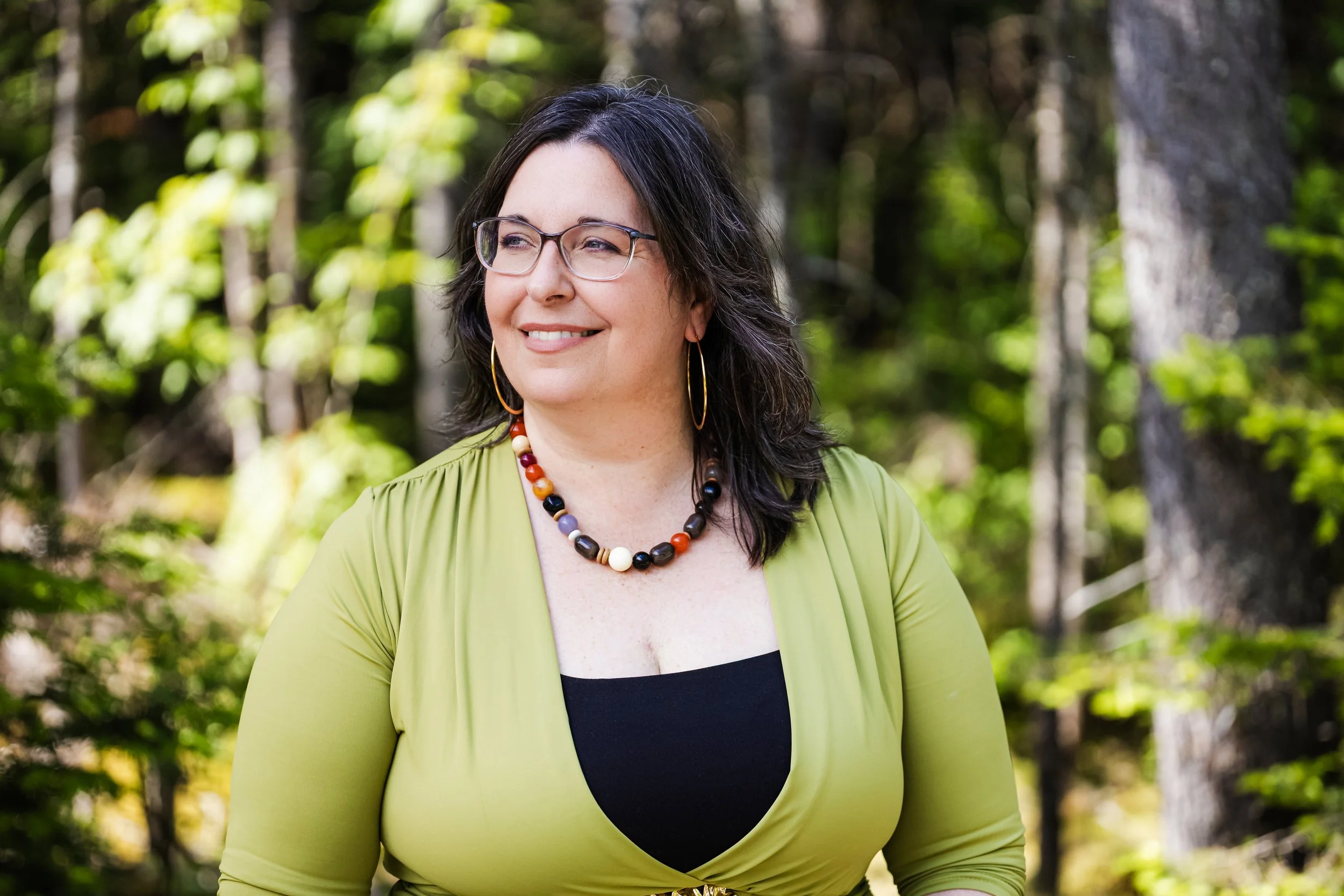 Une femme souriante porte des lunettes et un collier coloré, debout dans une forêt avec des arbres en arrière-plan.