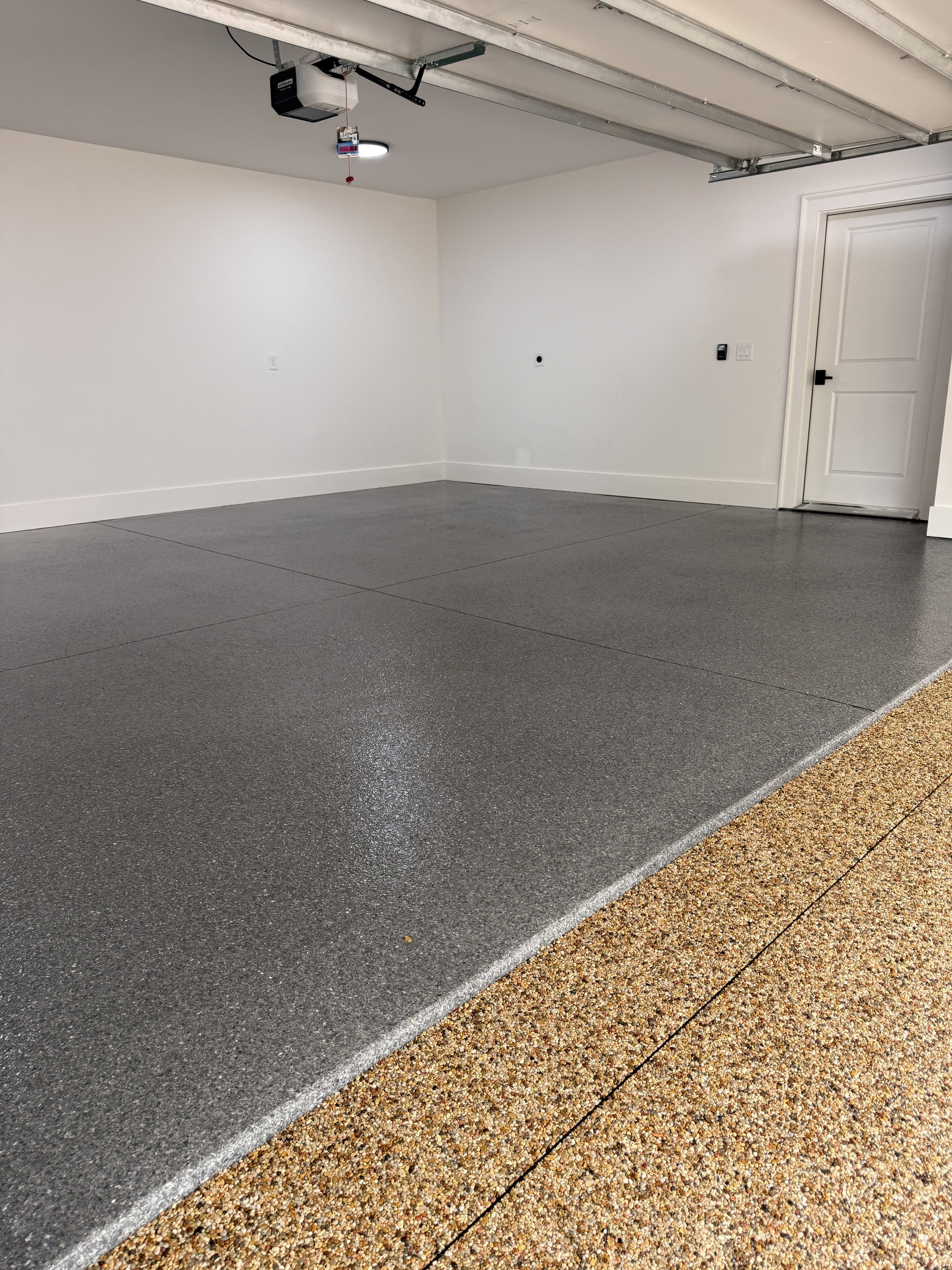 A garage with a resin-bound pebble garage and drivewadesigned and installed by Pebble & Resin Co. The garage door is open, and the interior walls are painted white. There is a door on the right side and a garage door opener is mounted on the ceiling.