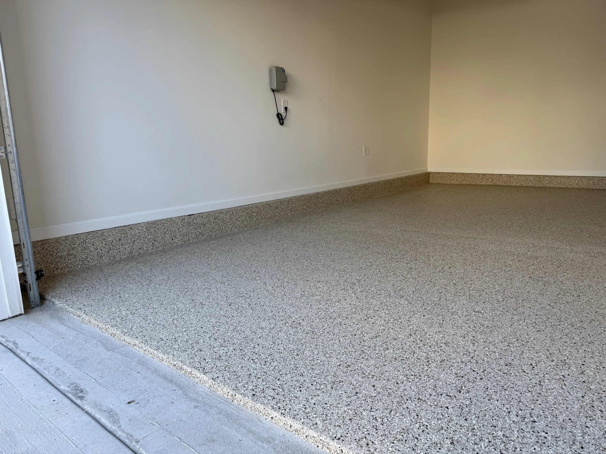 Empty garage with resin-bound pebble flooring designed and installed by Pebble & Resin Co, white walls, and a partially open garage door on the left side.