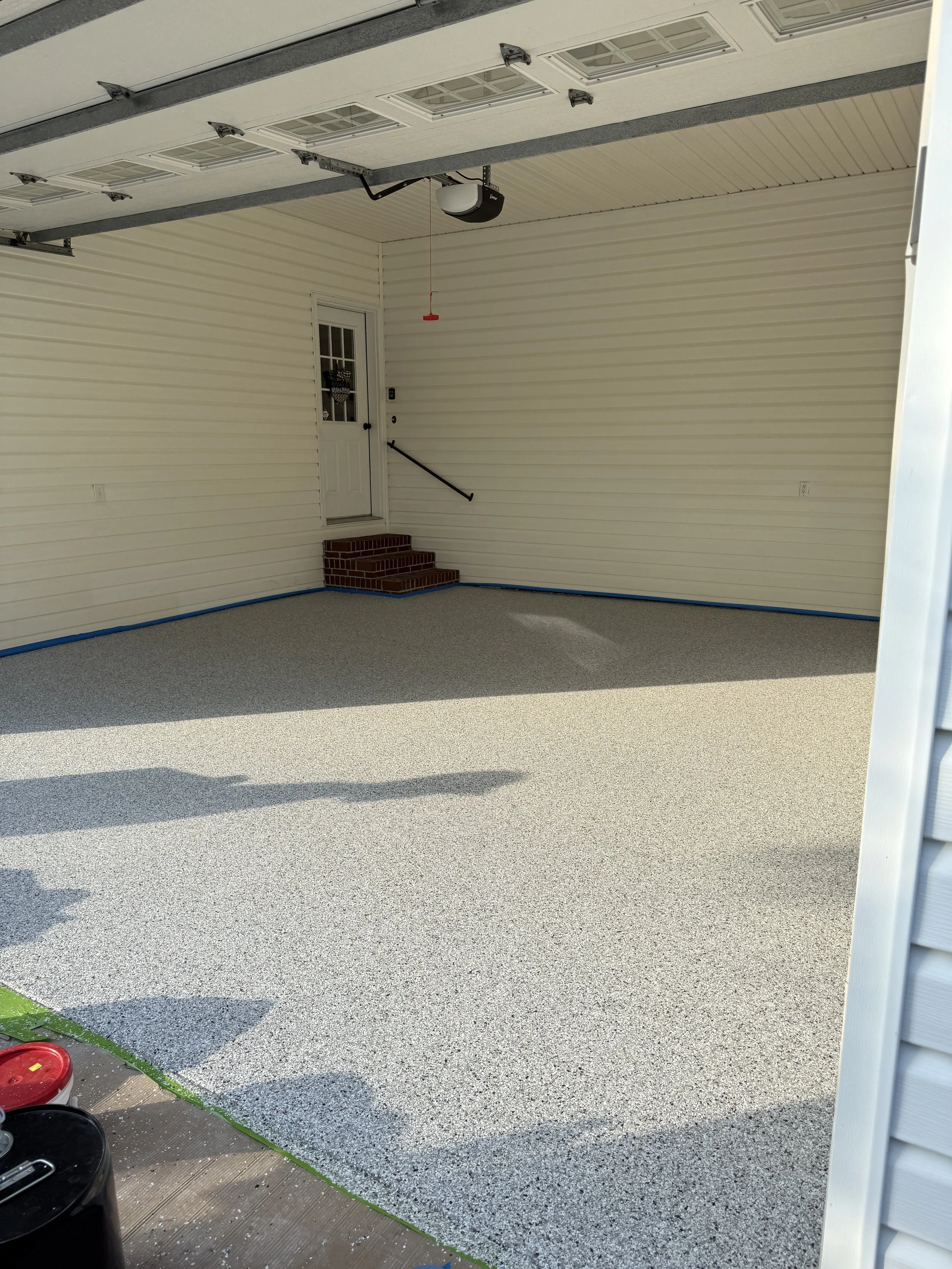 A newly poured pebble-gravel epoxy garage floor, beige in color, resin-bound pebble flooring designed and installed by Pebble & Resin Co with some tools and supplies visible in the corner.