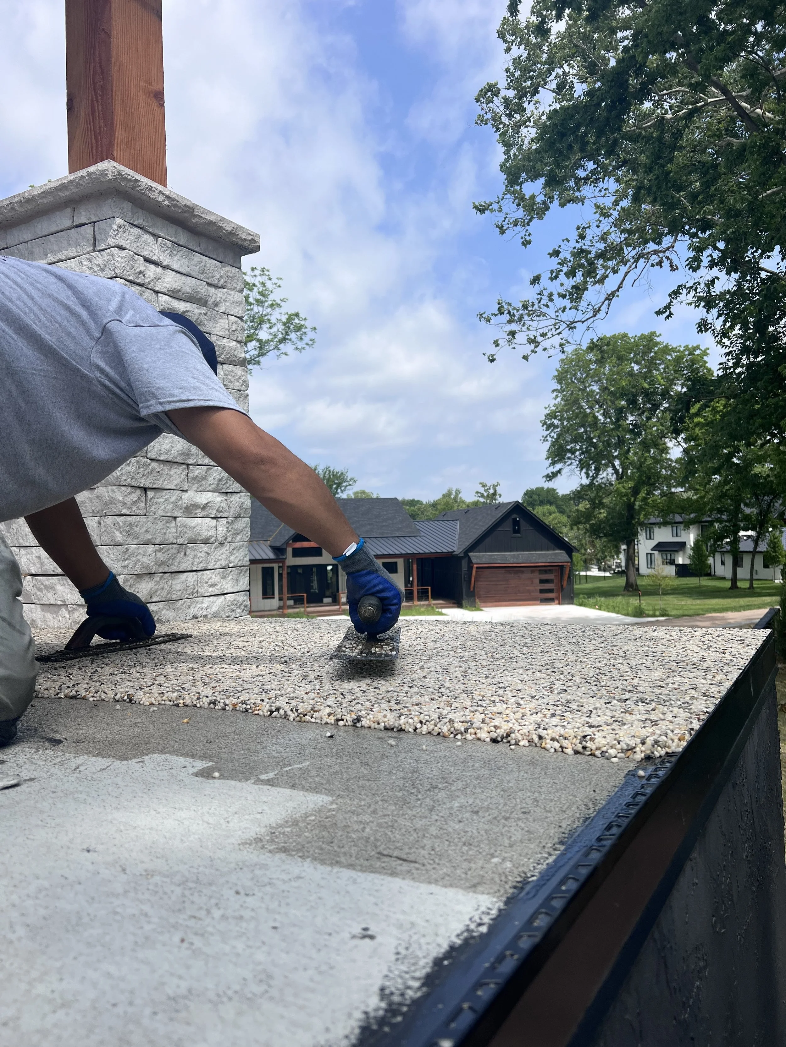 A person applying an outdoor epoxy coating on a flat surface with a roller, with a stone chimney and trees in a neighborhood background.