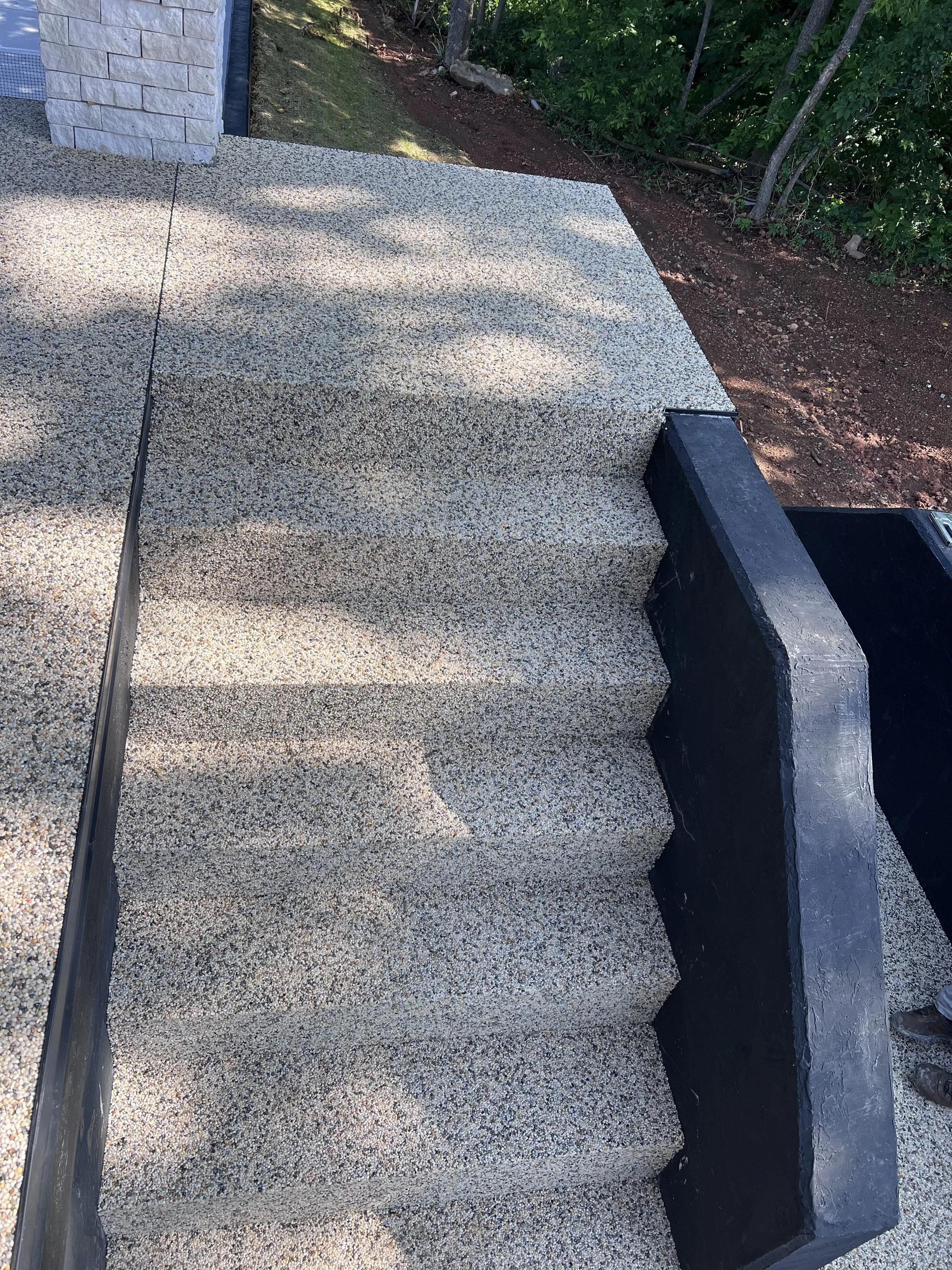 Close-up view of a newly installed Epoxy bound pebble stairs designed and installed by Pebble & Resin Co, with a dirt and grassy area and trees in the background.