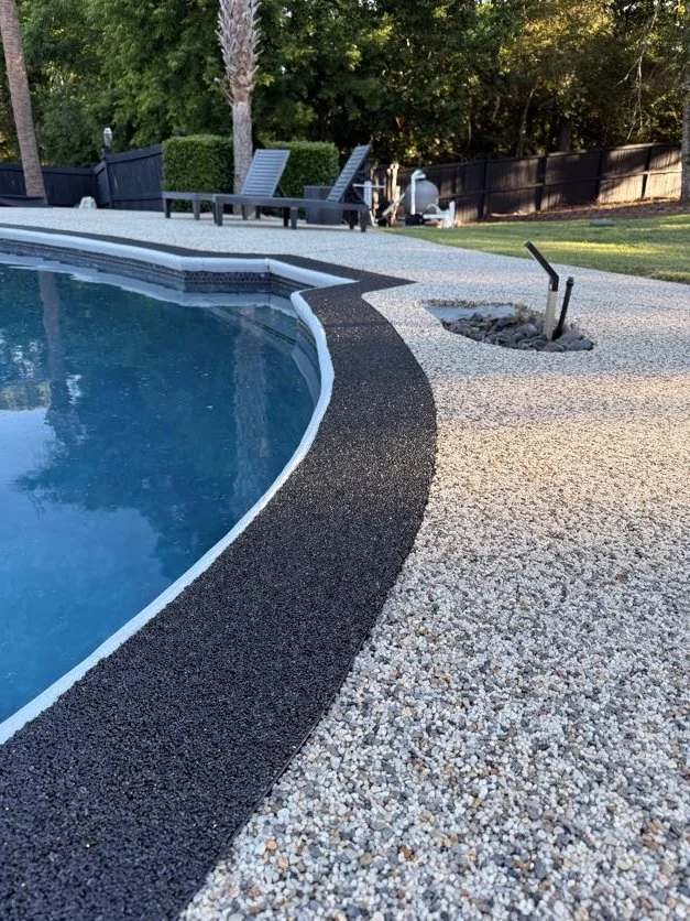Backyard swimming pool with resin-bound pebble pool deck designed and installed by Pebble & Resin Co black and white textured concrete pool deck, poolside lounge chairs, and a black fence with trees in the background.