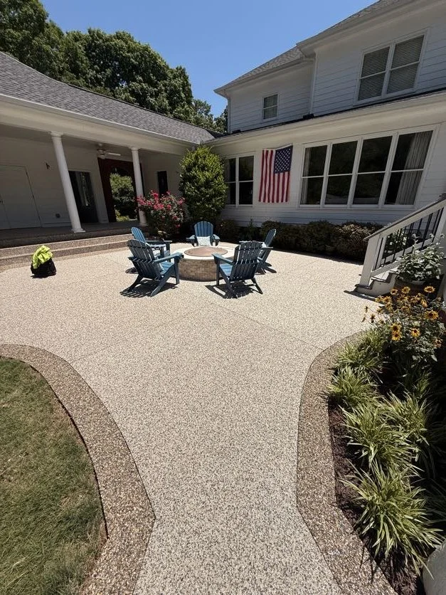 A backyard patio with blue Adirondack chairs around a fire pit, garden beds, a large American flag on the house, and a white house with a porch and stairs, under a clear blue sky.