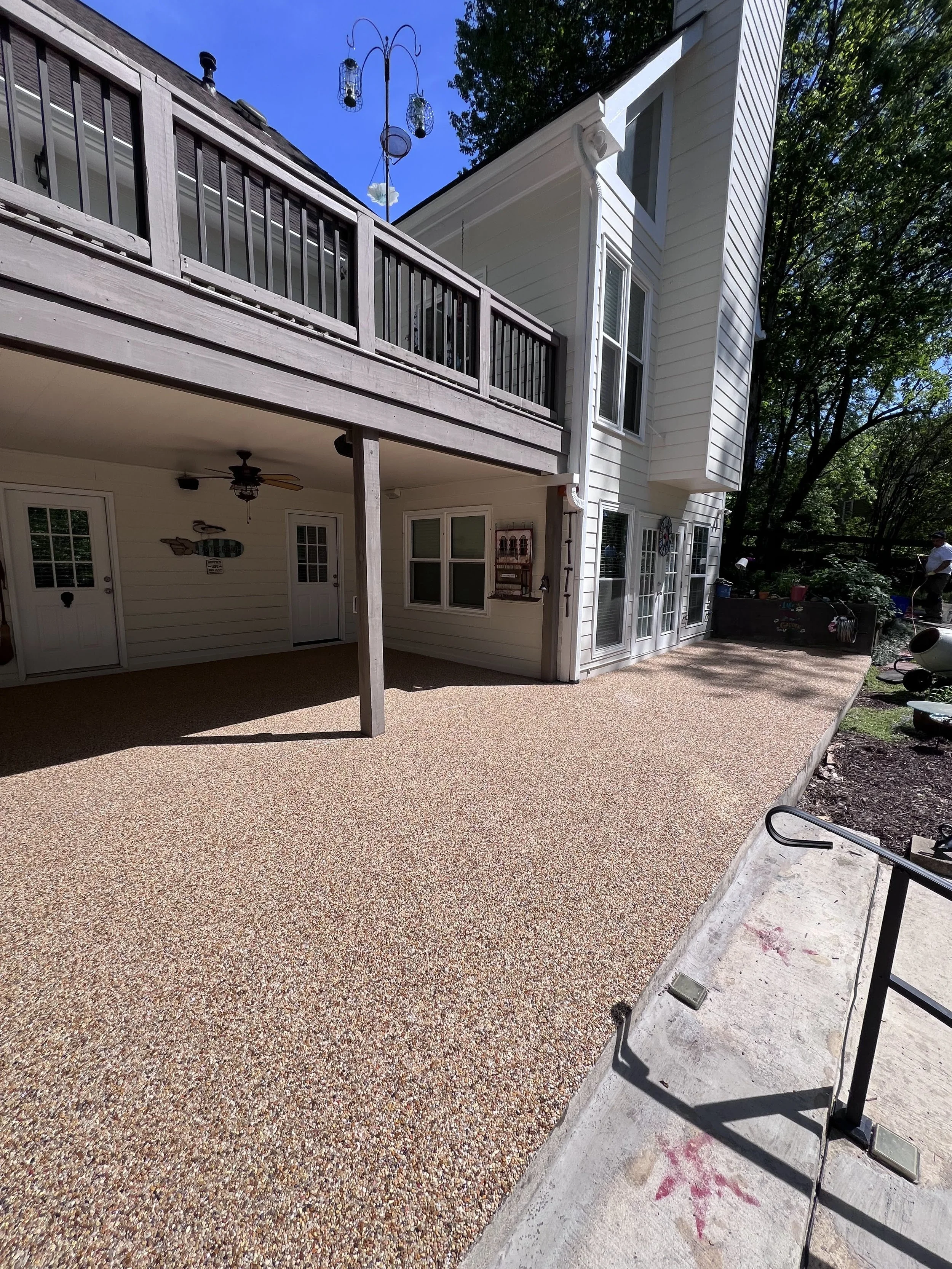 View of a house's back porch with a wooden deck, a ceiling fan, and several windows. There is a patio area with textured flooring, a small railing, and garden tools on the side. A man is seen in the background near the garden area.