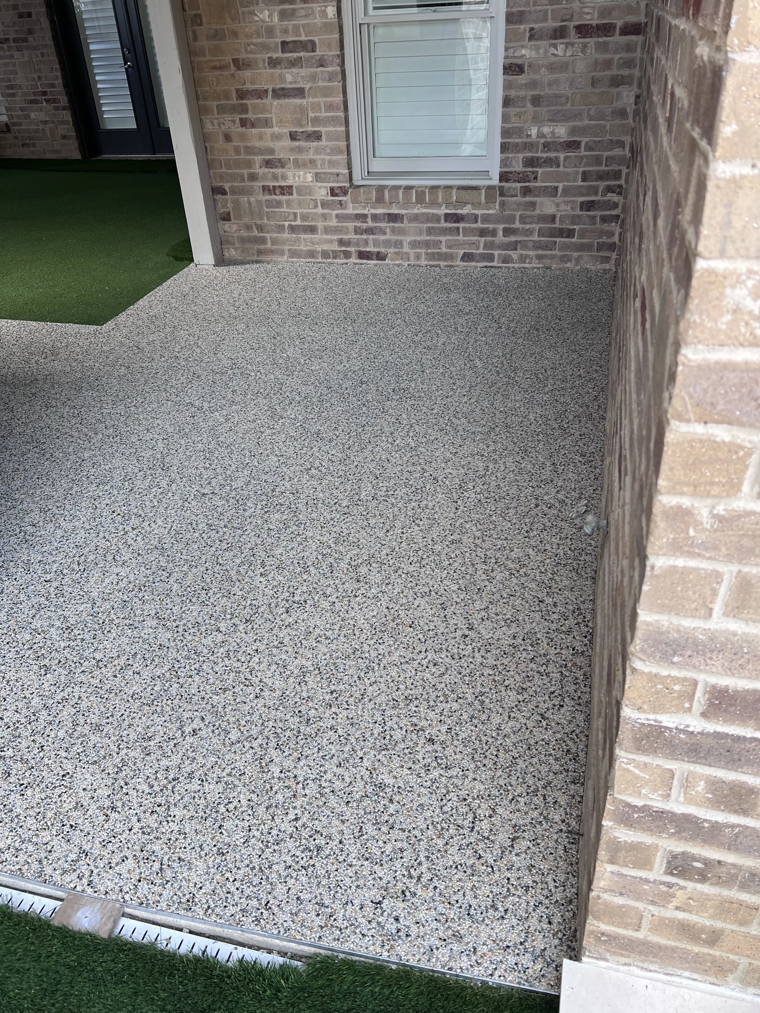 A small outdoor area with a synthetic grass strip, a gravel patio, and brick walls. There's a window with shutters on the brick wall, and a door with blinds visible. The area is bordered by a plastic or metal edging.