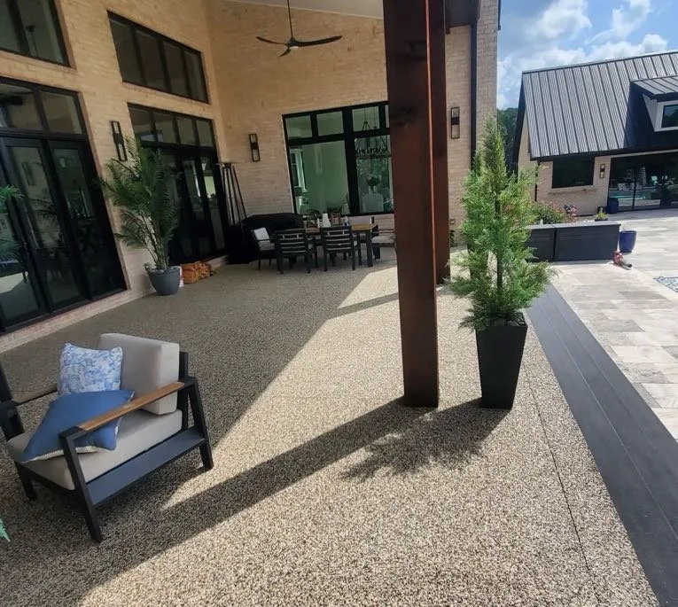 Outdoor patio with beige floor, potted plants, a beige armchair with a blue pillow, black chairs with a table, and a building with large glass windows and a grey roof.