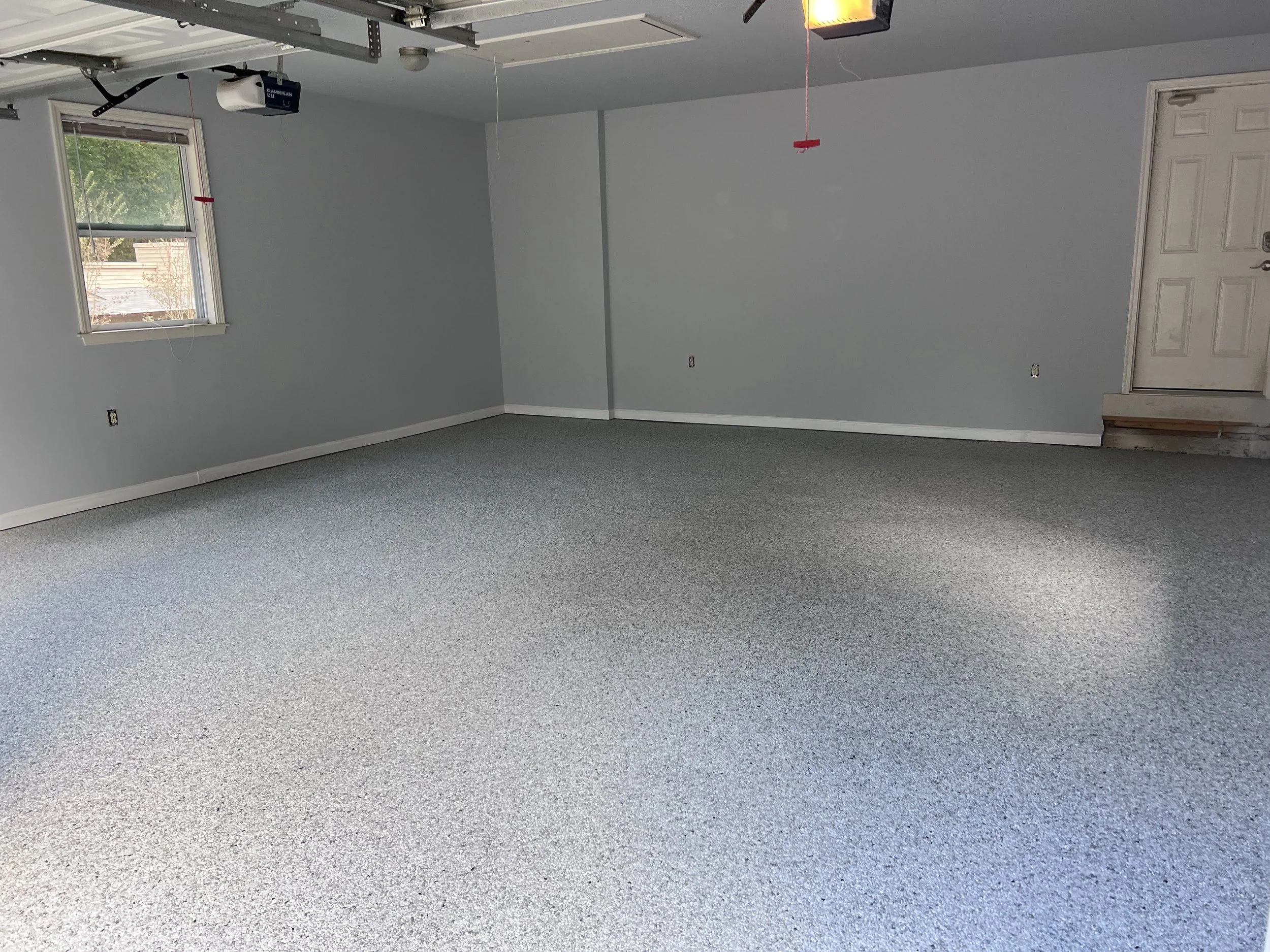 Empty garage with light grey walls, a window showing a tree outside, a closed door, and a resin-bound pebble flooring designed and installed by Pebble & Resin Co