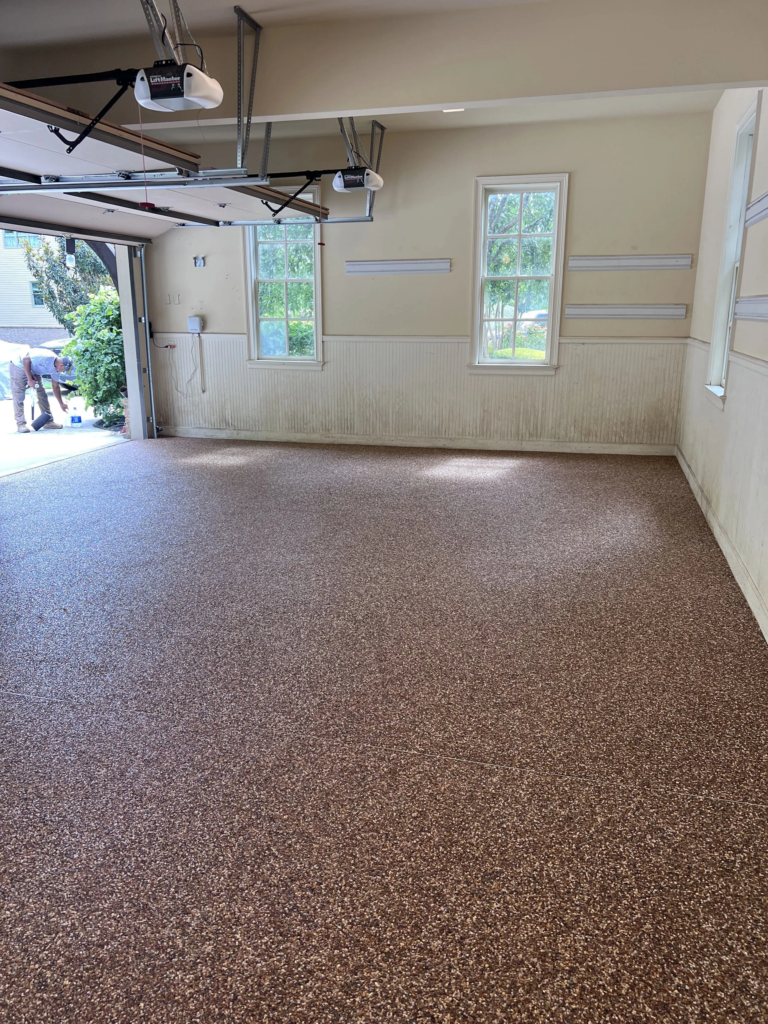 Empty garage with a resin-bound pebble flooring designed and installed by Pebble & Resin Co, two windows, and an open garage door. A person is outside near the garage, working with tools.
