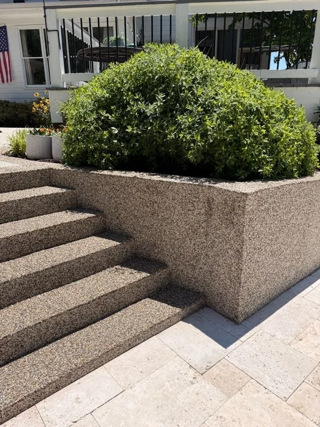 Epoxy bound pebble stairs designed and installed by Pebble & Resin Co steps leading up to a garden bed with a large green shrub in front of a house with white siding and black railing porch