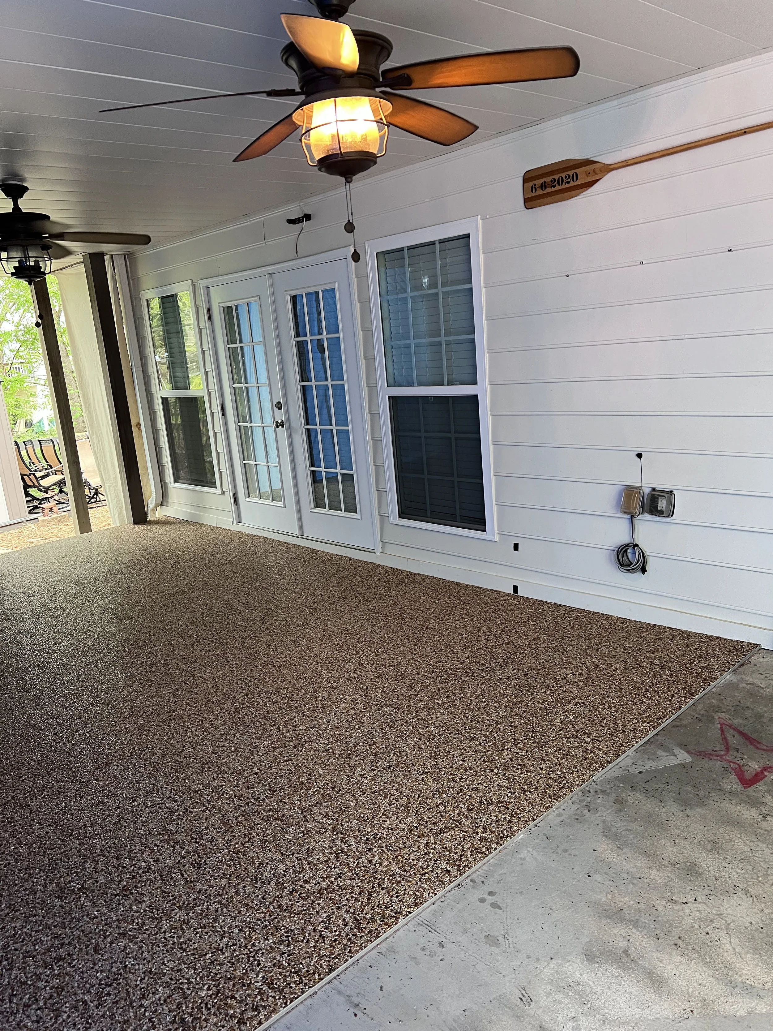 Exterior porch with two ceiling fans, one with a light turned on, white siding, two glass-paneled doors, and a window. The porch floor is covered with textured material, and there's a concrete section at the edge.