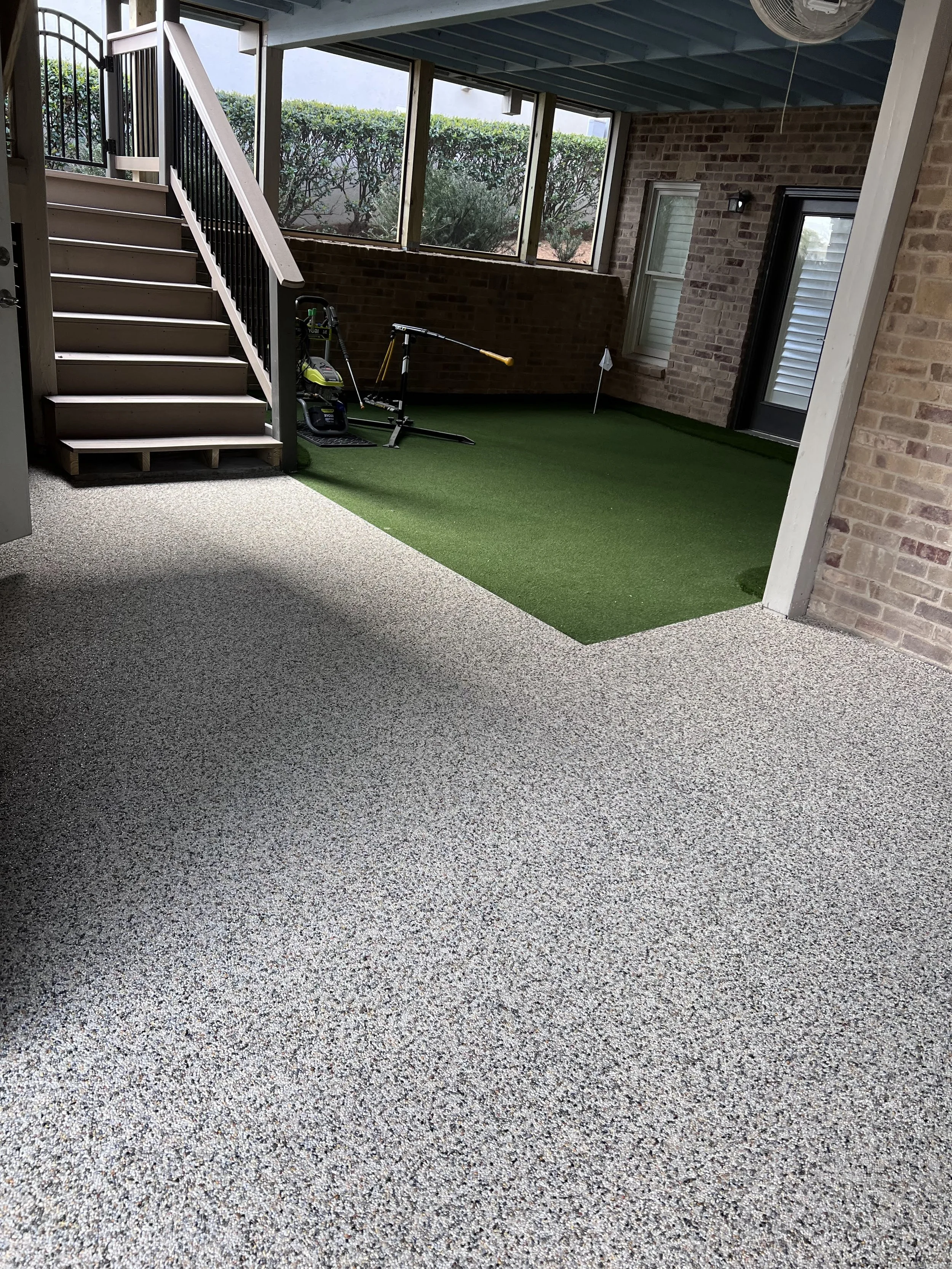 A covered patio area with a staircase, enclosed with screened windows, in a residential backyard. An artificial grass patch is on the floor with a golf club and a small black object nearby.