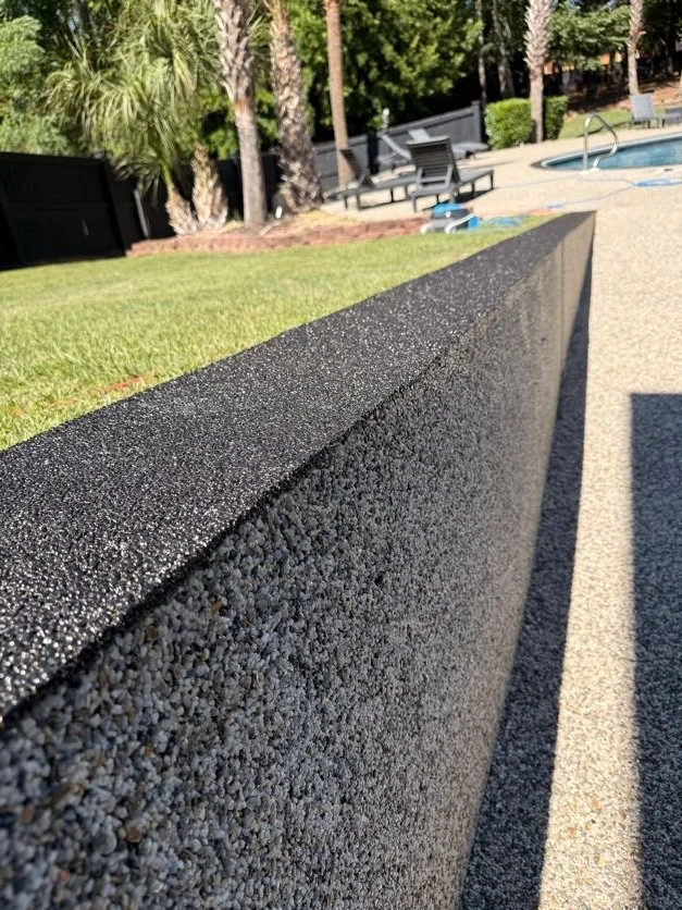 Close-up of a resin-bound pebble pool deck designed and installed by Pebble & Resin Co black textured wall with a grassy area and outdoor chairs and trees in the background.