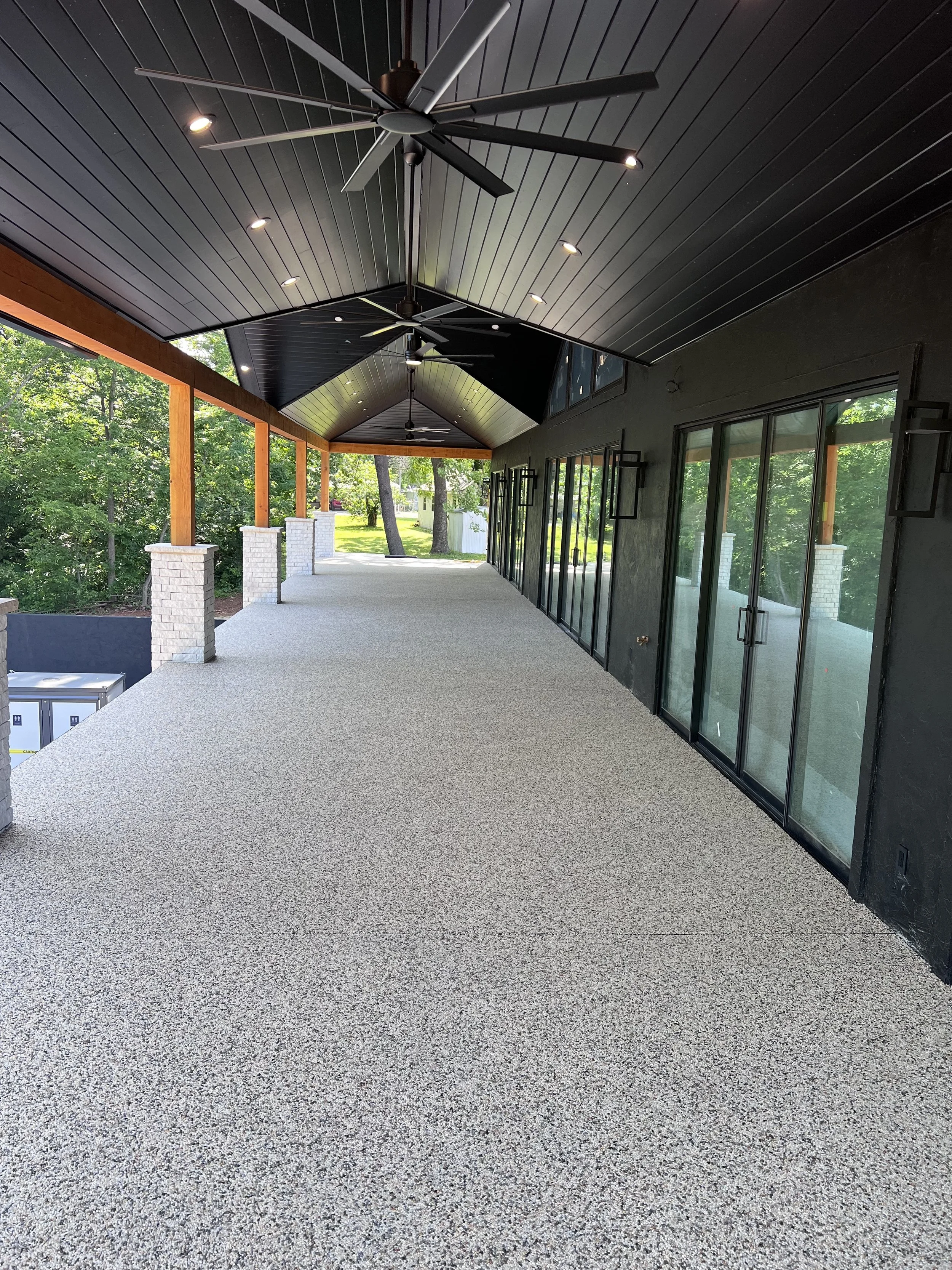 Covered patio with black ceiling, three ceiling fans, recessed lighting, large glass sliding doors, and a beige textured outdoor carpet. Surrounding green trees are visible through the open side.