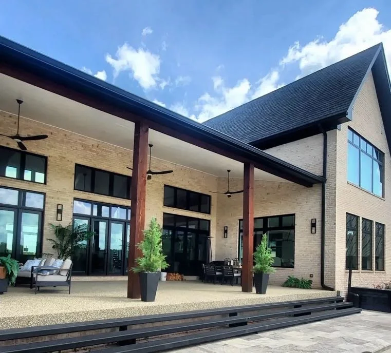 Large modern house with brick exterior, large windows, and a covered patio with outdoor furniture, potted plants, and ceiling fans, under a partly cloudy sky.