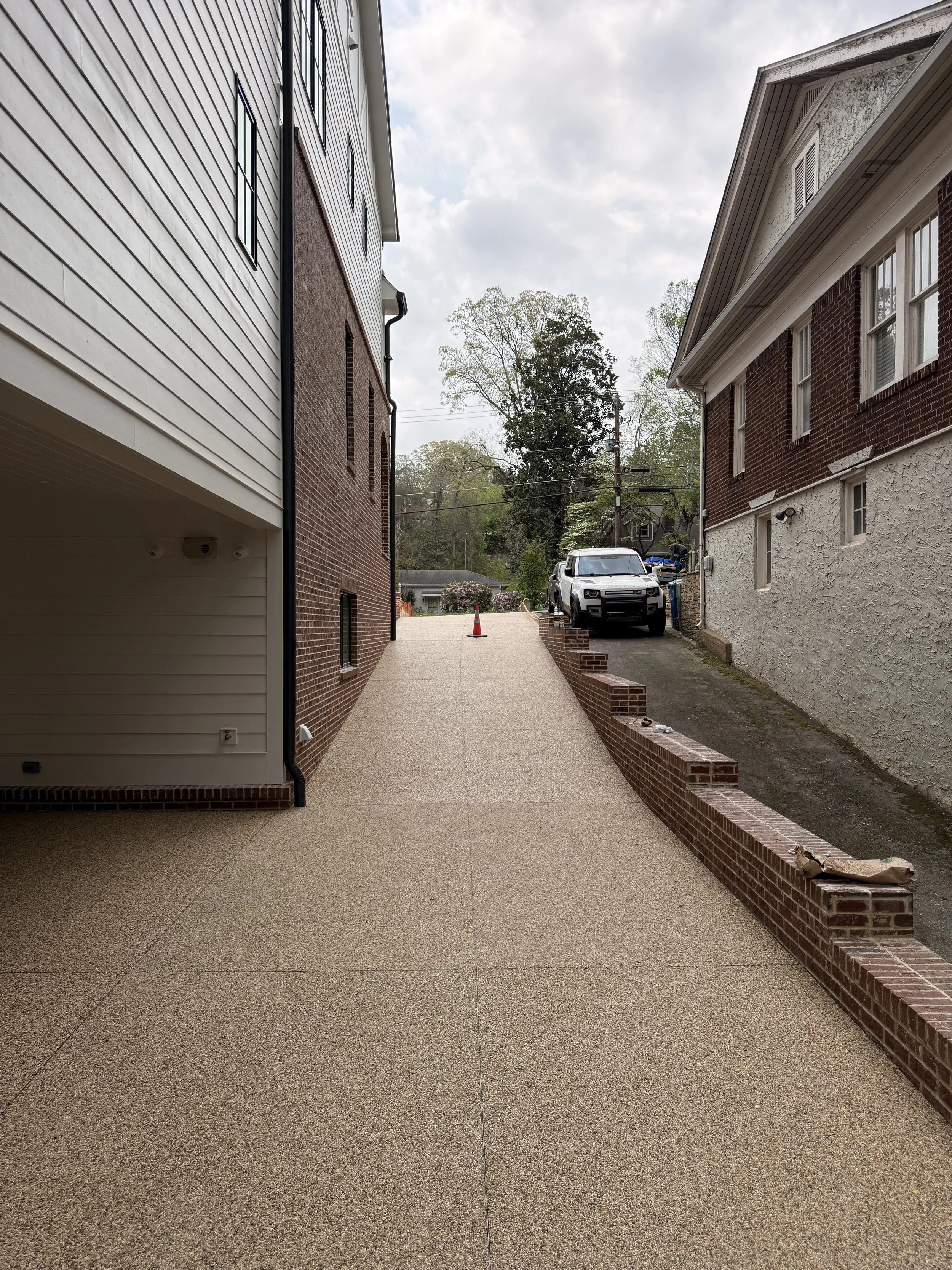 Residential resin-bound pebble driveway designed and installed by Pebble & Resin Co with brick and stucco houses, parked vehicle, street cones, and trees in the background under cloudy sky.