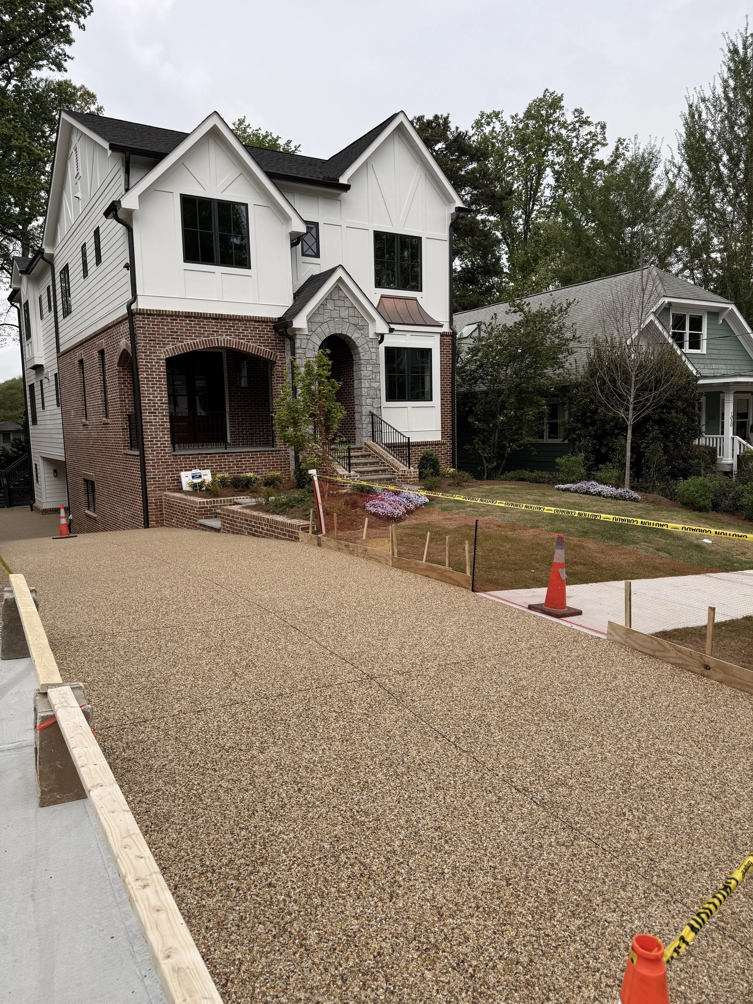 Photo of a newly paved driveway with resin-bound pebbles designed and installed by Pebble & Resin , orange safety cones, and caution tape, in front of two houses under construction or renovation, with landscaping and trees in the background.