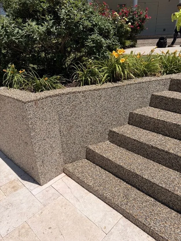 Epoxy bound pebble stairs designed and installed by Pebble & Resin Co next to a raised garden bed with green plants and yellow flowers, with more plants and a person in the background.