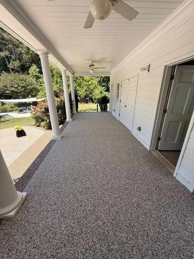 Long covered porch with Epoxy bound pebbled flooring designed and installed by Pebble & Resin Co, white siding, ceiling fans, and white columns, overlooking a garden with shrubs, flowers, and trees.