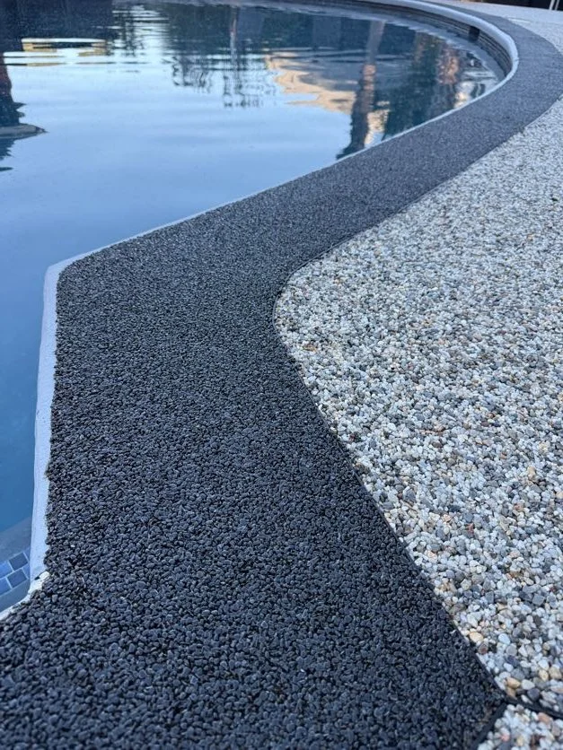 Close-up of a swimming pool resin-bound pebble pool deck designed and installed by Pebble & Resin Co edge with black and white textured surface materials, with the pool water visible in the background.