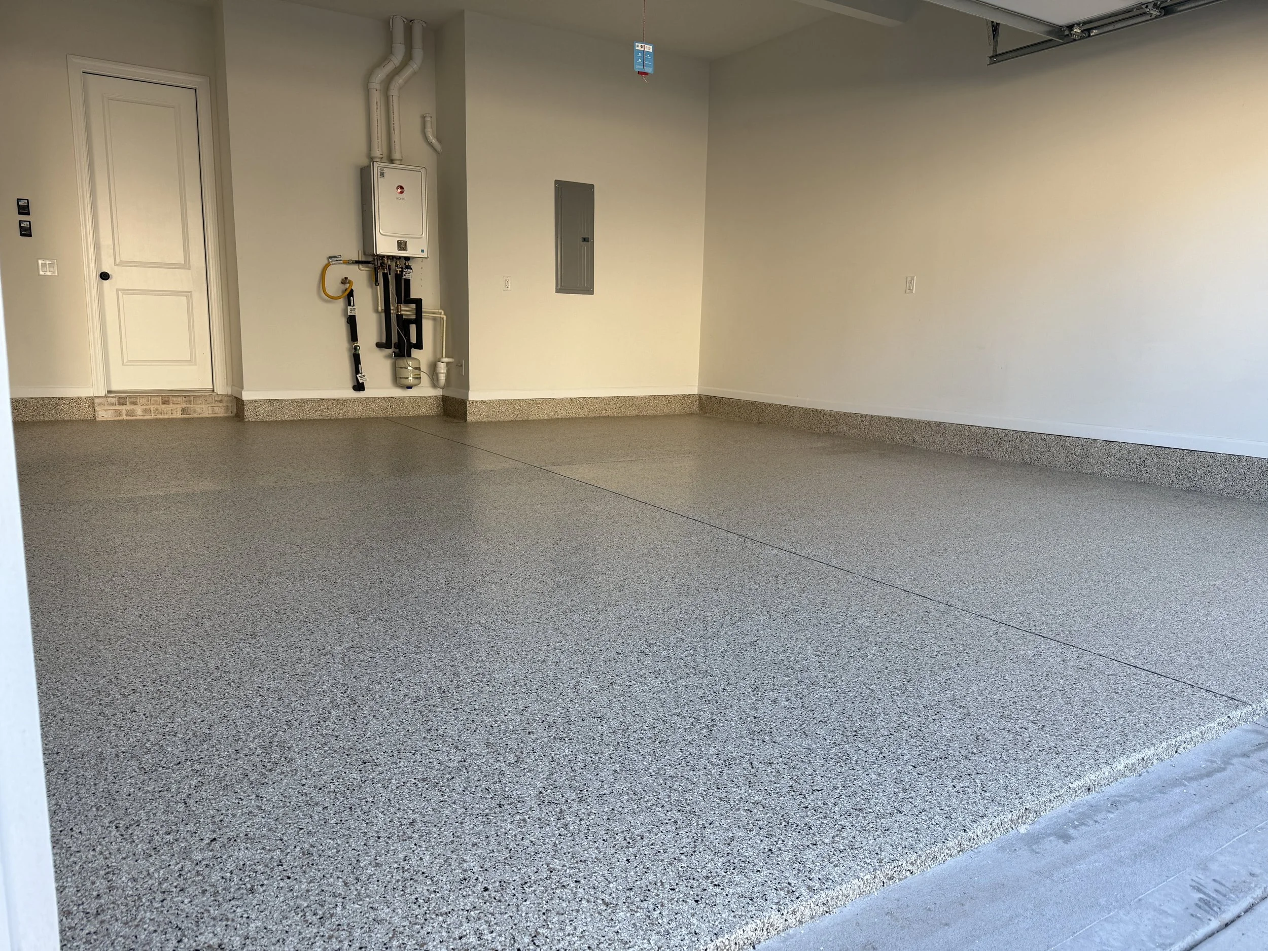 An empty garage with a speckled gray epoxy floor, white walls, a closed door on the left, a utility panel, and a garage door opener mechanism at the top right. resin-bound pebble flooring designed and installed by Pebble & Resin Co